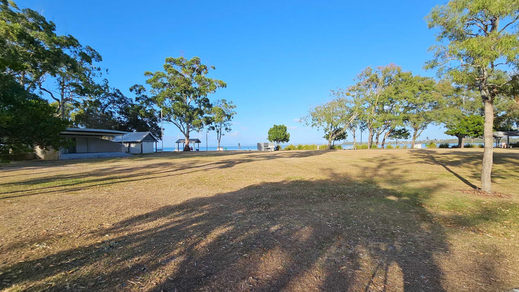 A grassy area with scattered trees casting long shadows. On the left, there is a small building with a sloped roof, partially obscured by trees. In the centre, a path leads towards the water, with a few people visible near a gazebo. On the right, more trees line the edge of the grass, with a glimpse of the sea in the background. The ground is a mix of dry grass and patches of dirt. A grassy area with scattered trees casting long shadows. On the left, there is a small building with a sloped roof, partially obscured by trees. In the centre, a path leads towards the water, with a few people visible near a gazebo. On the right, more trees line the edge of the grass, with a glimpse of the sea in the background. The ground is a mix of dry grass and patches of dirt.