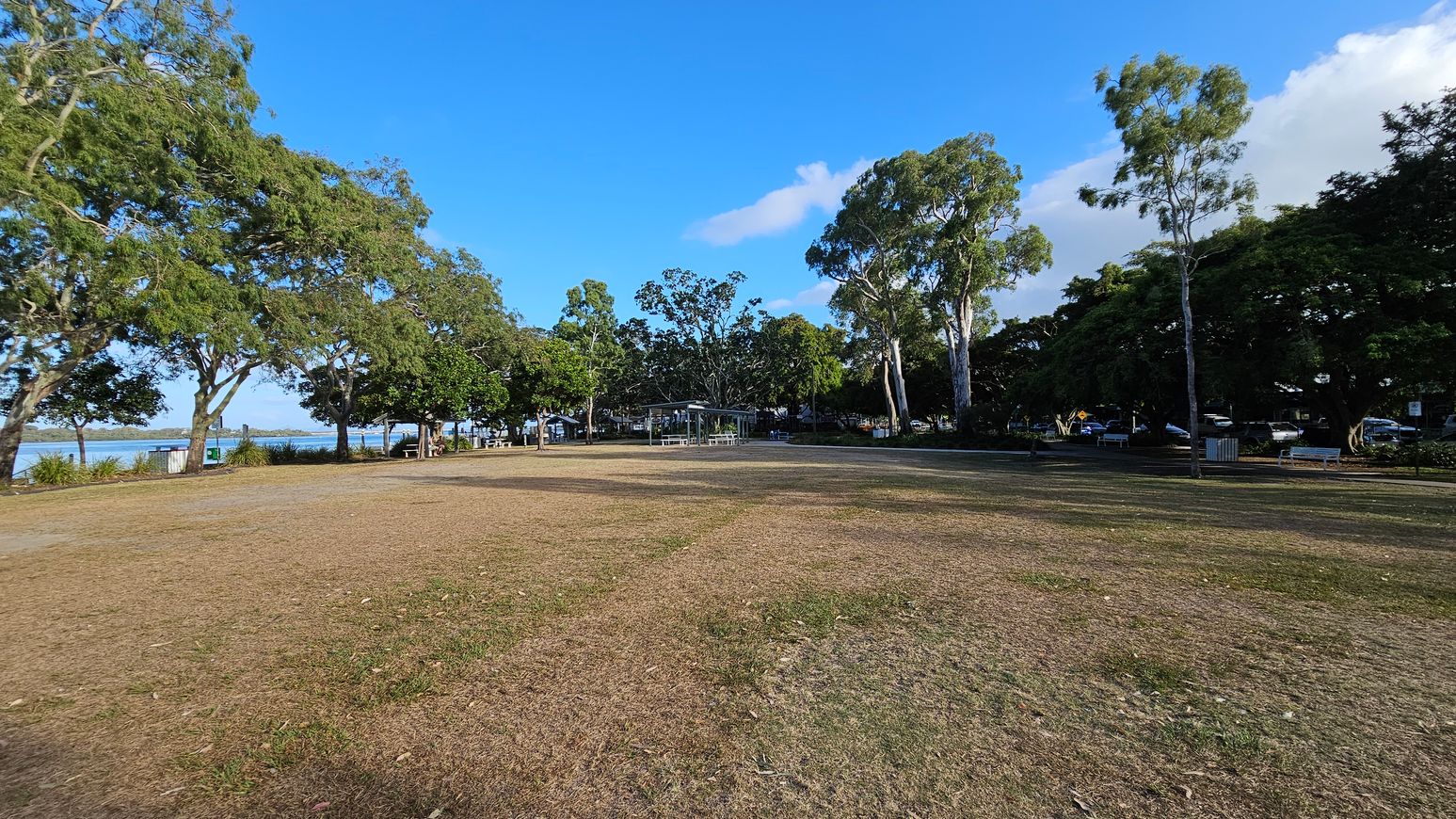 A grassy park area with patches of brown and green grass. On the left, there are large trees with dense foliage, and a glimpse of water is visible in the background. In the centre, a small pavilion with a metal roof is surrounded by more trees. To the right, additional trees and parked cars are visible, along with a few benches and a bin. The sky is clear and blue. A grassy park area with patches of brown and green grass. On the left, there are large trees with dense foliage, and a glimpse of water is visible in the background. In the centre, a small pavilion with a metal roof is surrounded by more trees. To the right, additional trees and parked cars are visible, along with a few benches and a bin. The sky is clear and blue.