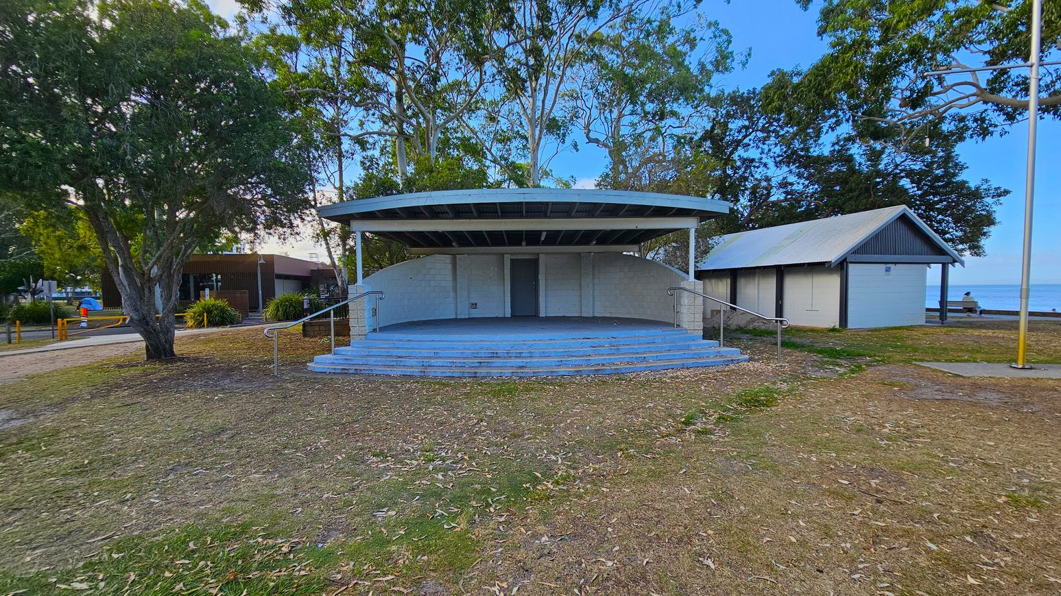 A small outdoor stage with a curved roof and steps leading up to it is in the centre. To the right, there's a small building with a sloped roof. On the left, a large tree provides shade, and a path leads to another structure in the background. The ground is covered with grass and scattered leaves, and a flagpole stands on the right. Trees are visible in the background, and the sea can be seen in the distance. A small outdoor stage with a curved roof and steps leading up to it is in the centre. To the right, there's a small building with a sloped roof. On the left, a large tree provides shade, and a path leads to another structure in the background. The ground is covered with grass and scattered leaves, and a flagpole stands on the right. Trees are visible in the background, and the sea can be seen in the distance.