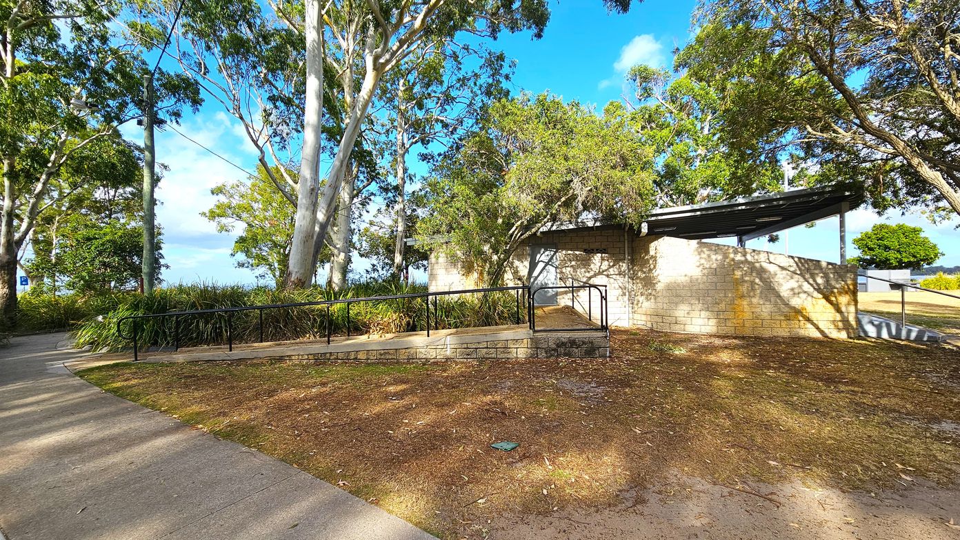 A paved path curves through a park area with tall trees and dense greenery on the left. In the centre, a brick structure with a sloped roof is partially obscured by trees. A metal railing runs alongside a ramp leading to the building. The ground is covered with dry leaves and grass. On the right, a small tree and open grassy area are visible. Shadows from the trees create patterns on the ground and building. A paved path curves through a park area with tall trees and dense greenery on the left. In the centre, a brick structure with a sloped roof is partially obscured by trees. A metal railing runs alongside a ramp leading to the building. The ground is covered with dry leaves and grass. On the right, a small tree and open grassy area are visible. Shadows from the trees create patterns on the ground and building.
