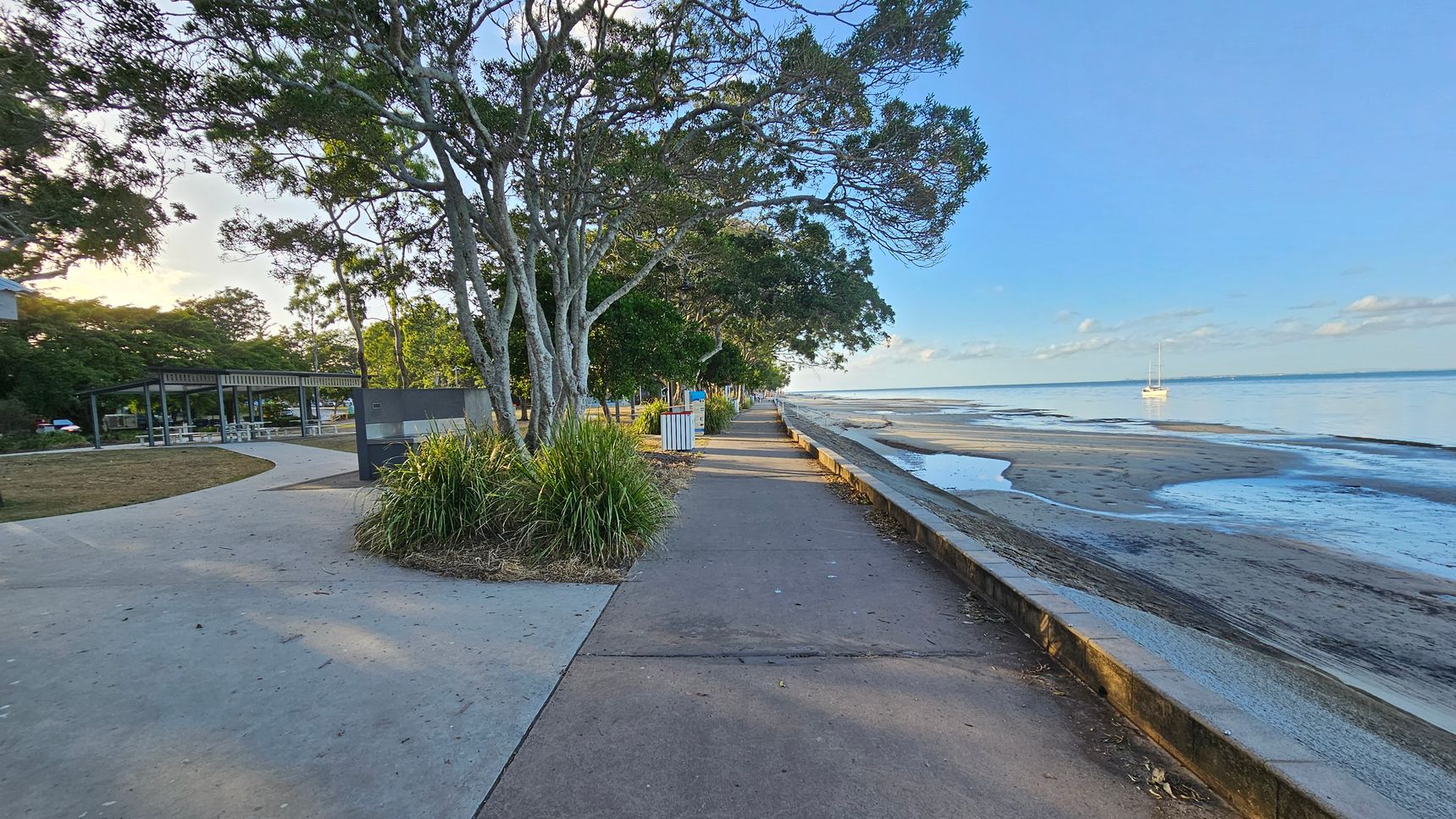 A coastal pathway with trees lining the left side, providing shade over a concrete walkway. To the left, there is a grassy area with a covered seating area and picnic tables. On the right, a low wall separates the path from a sandy beach with a calm sea. A sailboat is visible in the water. The sky is clear, and the area is well-lit, suggesting daytime. A coastal pathway with trees lining the left side, providing shade over a concrete walkway. To the left, there is a grassy area with a covered seating area and picnic tables. On the right, a low wall separates the path from a sandy beach with a calm sea. A sailboat is visible in the water. The sky is clear, and the area is well-lit, suggesting daytime.
