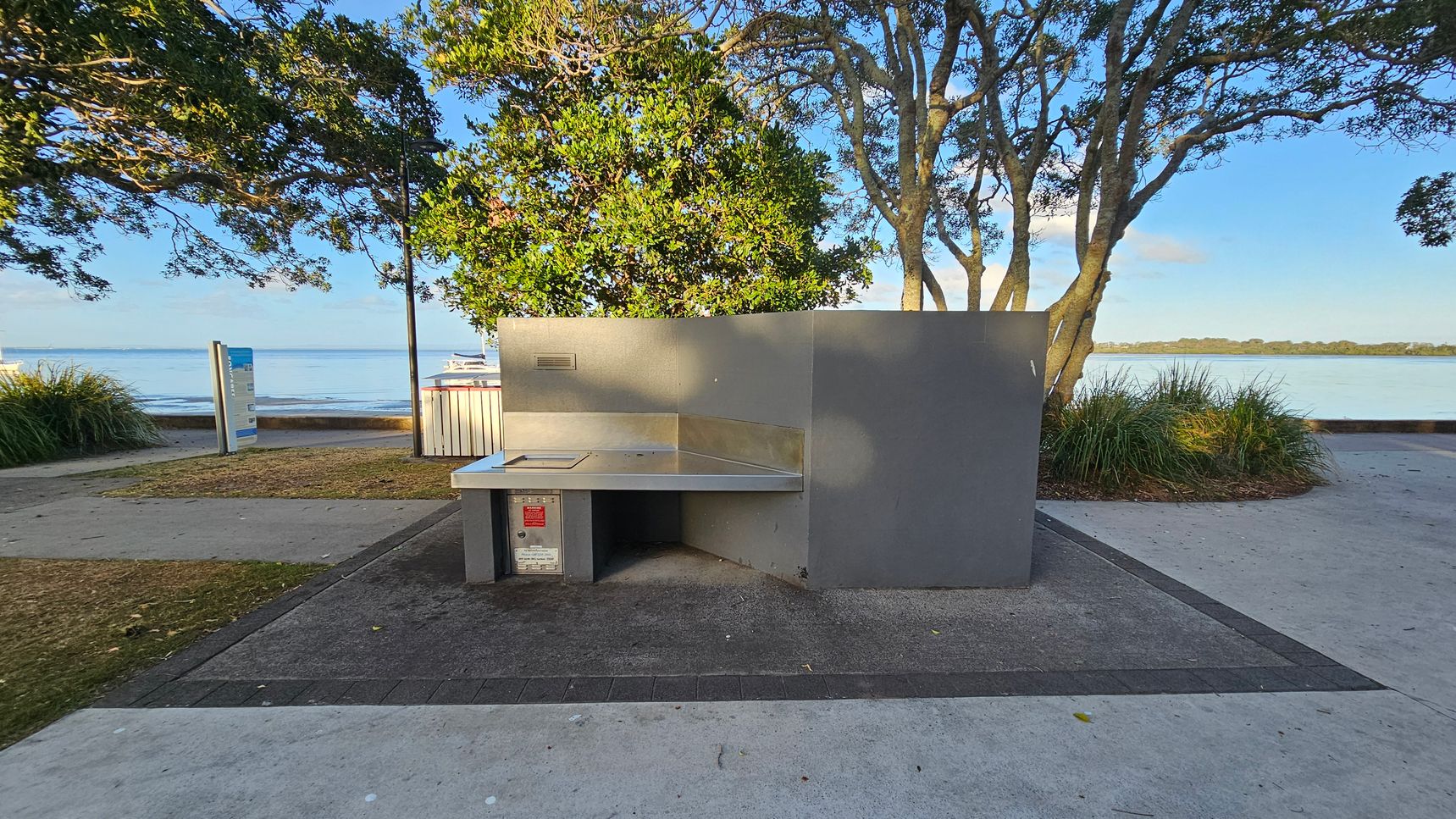 A public barbecue area with a stainless steel cooking surface is in the foreground, set on a paved area. To the left, a path leads towards a sign and a grassy patch. In the background, there are trees with dense foliage and a view of a calm body of water. A few boats are visible on the water, and there is a low wall separating the path from the water's edge. The scene is well-lit, suggesting daylight. A public barbecue area with a stainless steel cooking surface is in the foreground, set on a paved area. To the left, a path leads towards a sign and a grassy patch. In the background, there are trees with dense foliage and a view of a calm body of water. A few boats are visible on the water, and there is a low wall separating the path from the water's edge. The scene is well-lit, suggesting daylight.