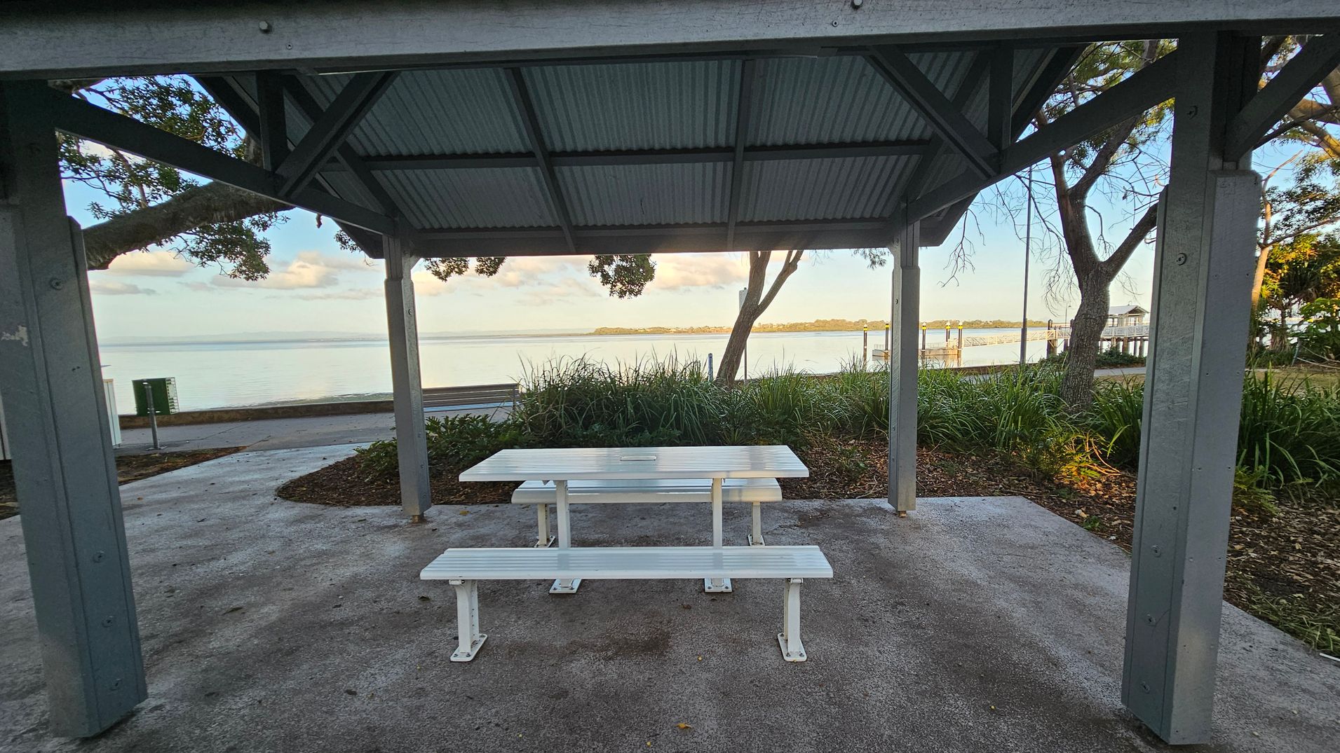 A sheltered picnic area with a white table and benches on a concrete floor. The structure has a corrugated metal roof supported by wooden beams. In the background, there is a view of a calm body of water with a distant shoreline. Trees and greenery surround the area, and a pathway runs parallel to the water. A small pier extends into the water on the right side. A sheltered picnic area with a white table and benches on a concrete floor. The structure has a corrugated metal roof supported by wooden beams. In the background, there is a view of a calm body of water with a distant shoreline. Trees and greenery surround the area, and a pathway runs parallel to the water. A small pier extends into the water on the right side.