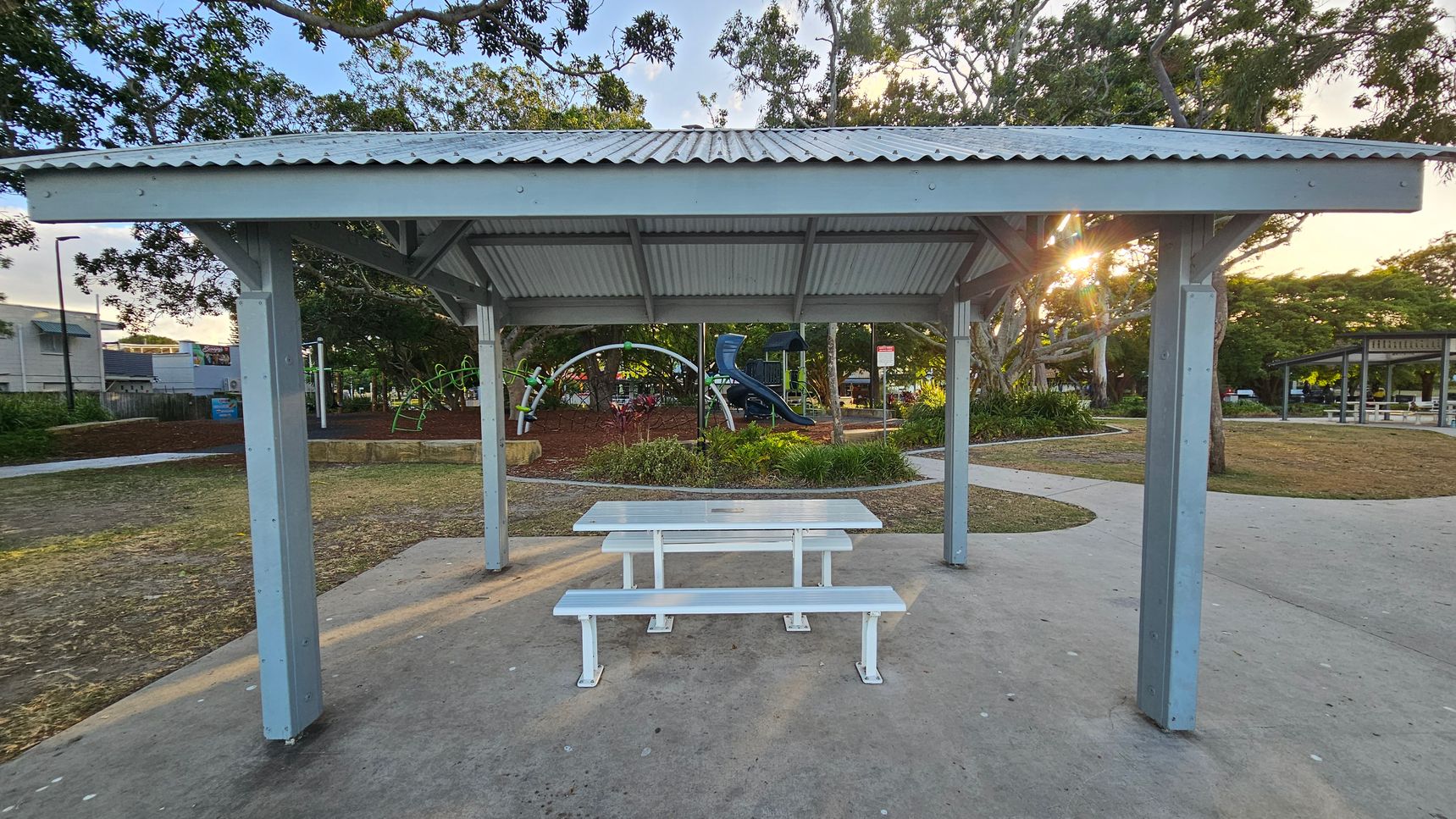 A park setting with a metal shelter in the foreground, featuring a corrugated roof and supported by four posts. Underneath is a white picnic table with benches. In the background, a playground with climbing structures and a slide is visible, surrounded by trees and greenery. A path curves around the area, and another shelter is seen to the right. The ground is a mix of concrete and grass, with some mulch around the playground. Sunlight filters through the trees. A park setting with a metal shelter in the foreground, featuring a corrugated roof and supported by four posts. Underneath is a white picnic table with benches. In the background, a playground with climbing structures and a slide is visible, surrounded by trees and greenery. A path curves around the area, and another shelter is seen to the right. The ground is a mix of concrete and grass, with some mulch around the playground. Sunlight filters through the trees.