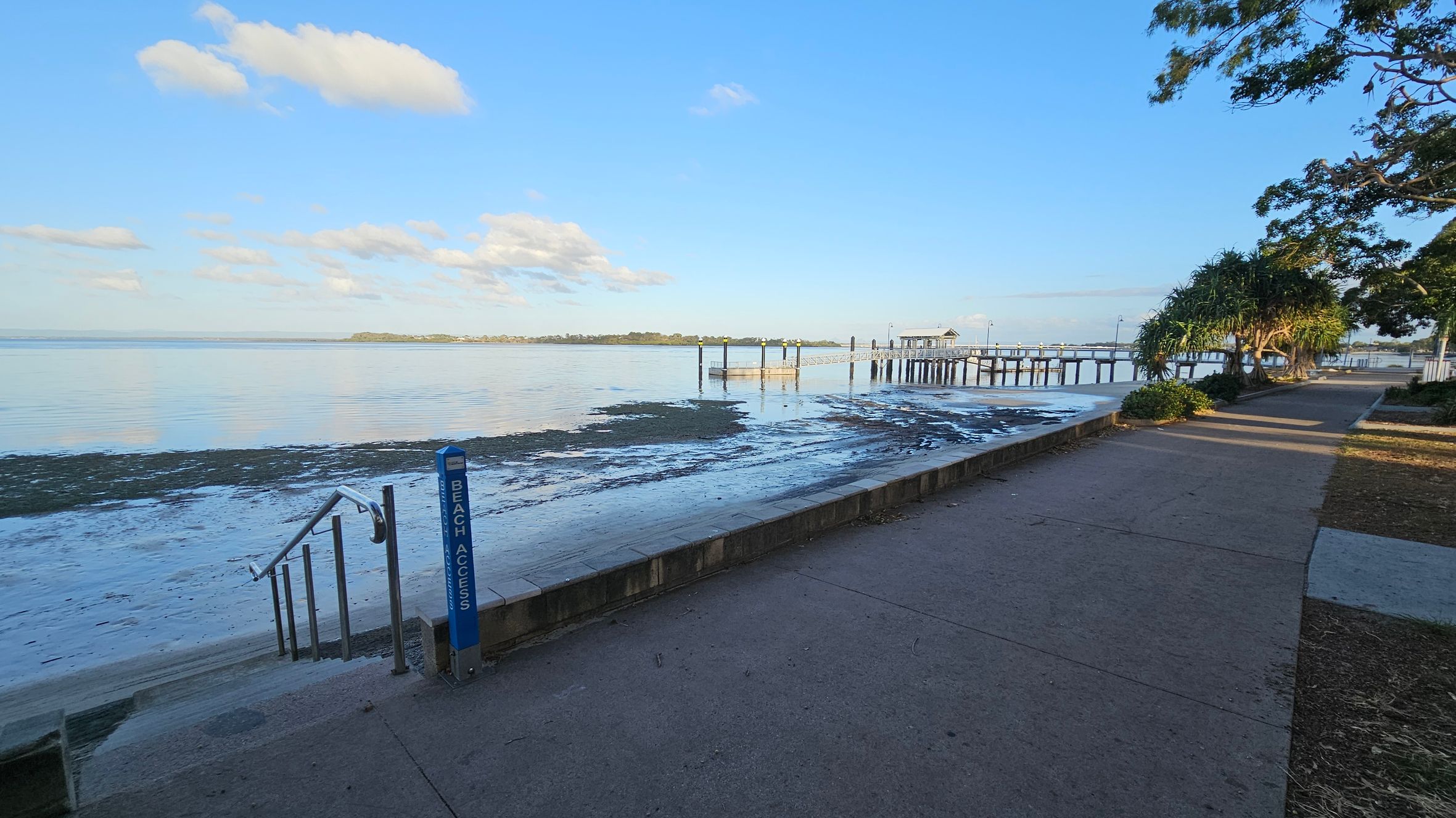 A coastal scene with a paved walkway on the right, bordered by trees and shrubs. A "Beach Access" sign and steps lead down to a sandy and muddy shoreline on the left. In the middle distance, a long pier extends over calm water, with a small structure at the end. The background features a distant landmass across the water. The sky is clear with a few clouds. A coastal scene with a paved walkway on the right, bordered by trees and shrubs. A "Beach Access" sign and steps lead down to a sandy and muddy shoreline on the left. In the middle distance, a long pier extends over calm water, with a small structure at the end. The background features a distant landmass across the water. The sky is clear with a few clouds.