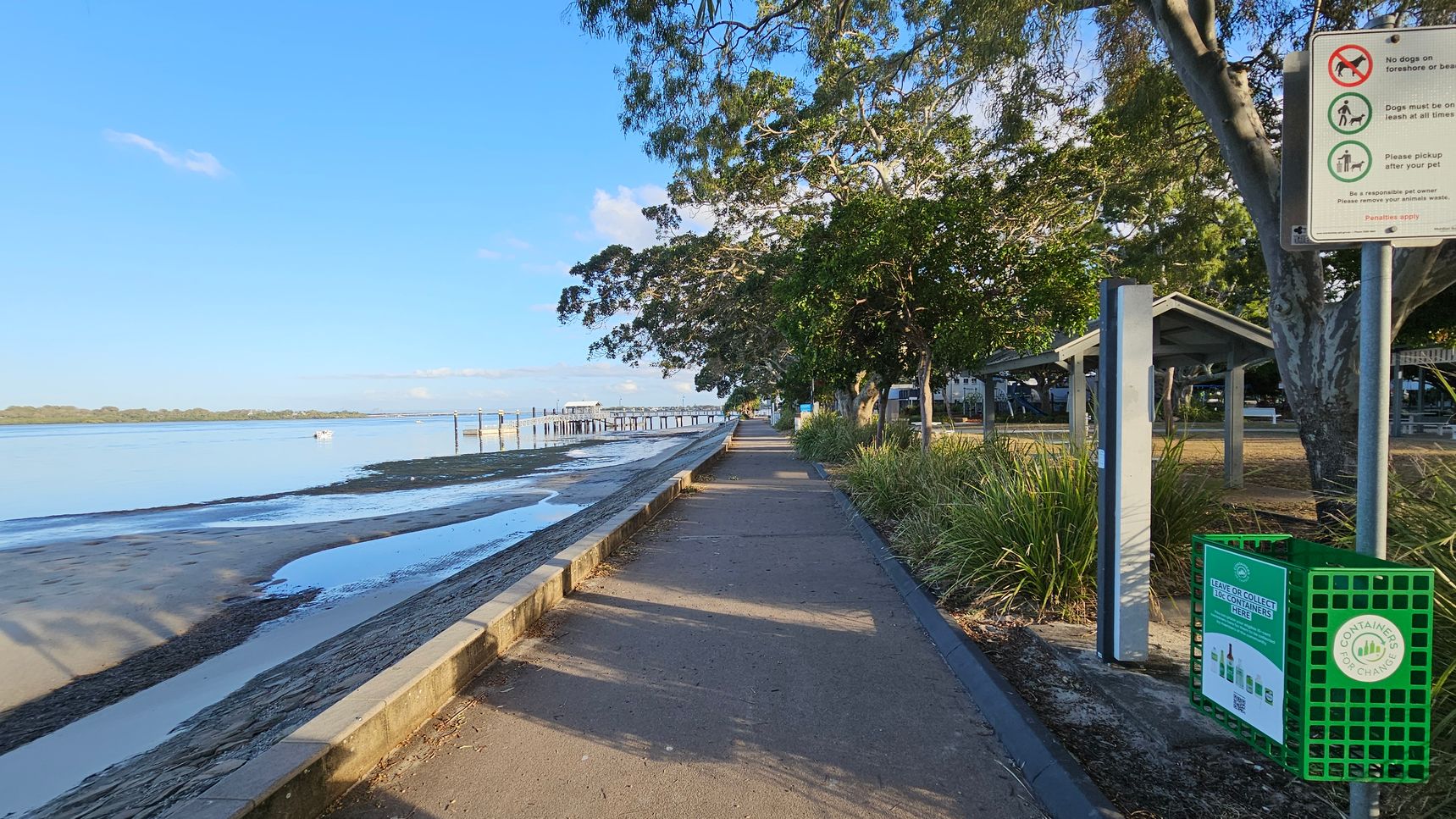A coastal pathway runs alongside a sandy beach on the left, with a low stone wall separating them. The beach leads to calm water with a distant pier. On the right, a signpost with pet-related rules stands near a green recycling bin. Trees provide shade over the path, and a small shelter with a sloped roof is visible among the greenery. The path is bordered by grass and shrubs, creating a serene, natural setting. A coastal pathway runs alongside a sandy beach on the left, with a low stone wall separating them. The beach leads to calm water with a distant pier. On the right, a signpost with pet-related rules stands near a green recycling bin. Trees provide shade over the path, and a small shelter with a sloped roof is visible among the greenery. The path is bordered by grass and shrubs, creating a serene, natural setting.