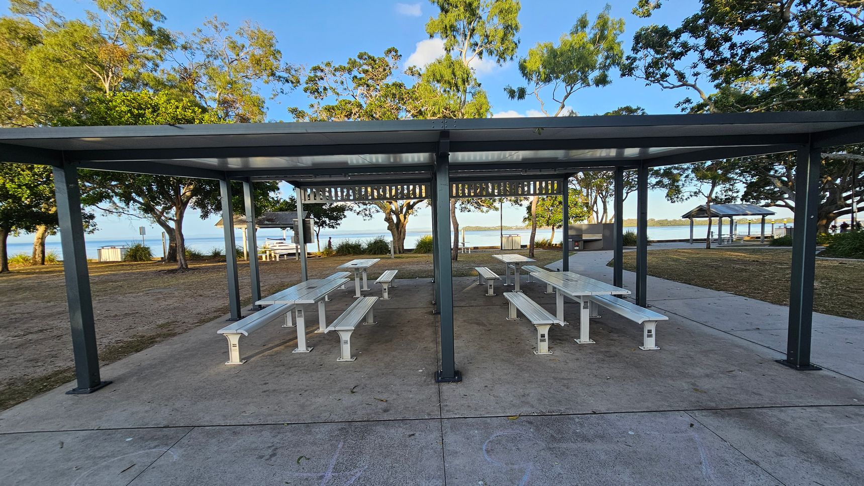 A covered picnic area with several white benches and tables on a concrete surface. The structure has a metal roof supported by dark green posts. In the background, there are trees with green foliage and a view of a body of water. To the right, a pathway leads to another covered area. The ground is a mix of grass and concrete, and the sky is clear. A covered picnic area with several white benches and tables on a concrete surface. The structure has a metal roof supported by dark green posts. In the background, there are trees with green foliage and a view of a body of water. To the right, a pathway leads to another covered area. The ground is a mix of grass and concrete, and the sky is clear.