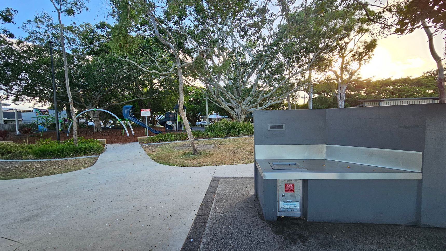 A park scene with a grey barbecue station on the right. In the middle, a paved path leads towards a playground area with climbing structures and slides. The left side features green grass and a variety of trees, including a large one with sprawling branches. The background is filled with more trees and a hint of a building. The ground is a mix of grass and mulch, and the sky is partially visible with a warm glow from the setting sun. A park scene with a grey barbecue station on the right. In the middle, a paved path leads towards a playground area with climbing structures and slides. The left side features green grass and a variety of trees, including a large one with sprawling branches. The background is filled with more trees and a hint of a building. The ground is a mix of grass and mulch, and the sky is partially visible with a warm glow from the setting sun.
