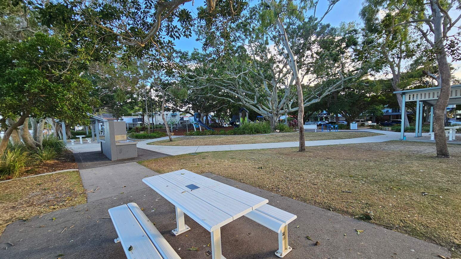 A park with a white picnic table and bench in the foreground on a paved path. To the left, there are trees and a small structure with a barbecue area. In the middle, a playground with a slide is visible among more trees. The background features additional seating areas and a few buildings partially obscured by foliage. The ground is a mix of grass and mulch, and the area is shaded by large trees. A park with a white picnic table and bench in the foreground on a paved path. To the left, there are trees and a small structure with a barbecue area. In the middle, a playground with a slide is visible among more trees. The background features additional seating areas and a few buildings partially obscured by foliage. The ground is a mix of grass and mulch, and the area is shaded by large trees.