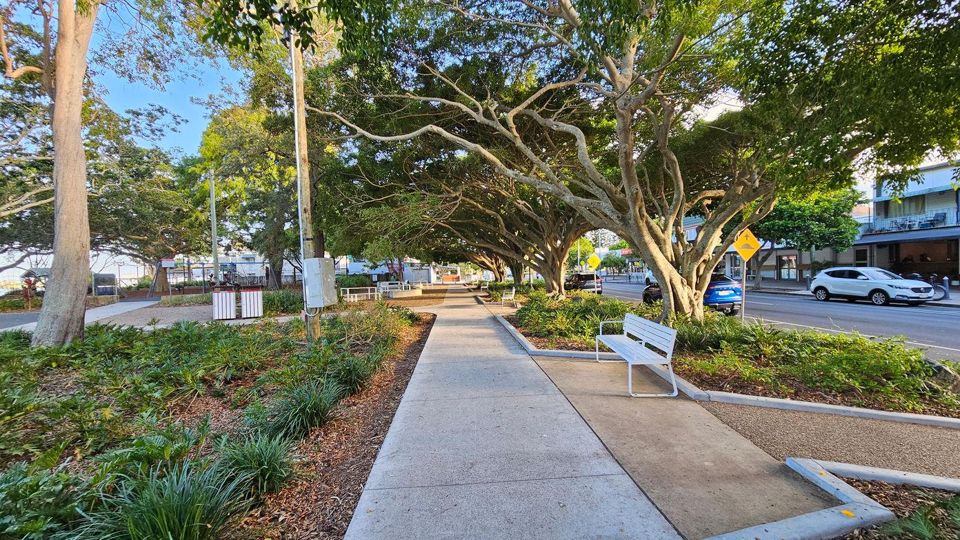 A pathway lined with white benches runs through a park area. On the left, there are large trees with thick trunks and lush green foliage, along with a grassy area and a utility pole. On the right, more trees provide shade, and a road with parked cars is visible. A yellow pedestrian crossing sign is near the road. The ground is a mix of concrete and mulch, with various plants and shrubs along the path. Buildings are visible in the background across the street. A pathway lined with white benches runs through a park area. On the left, there are large trees with thick trunks and lush green foliage, along with a grassy area and a utility pole. On the right, more trees provide shade, and a road with parked cars is visible. A yellow pedestrian crossing sign is near the road. The ground is a mix of concrete and mulch, with various plants and shrubs along the path. Buildings are visible in the background across the street.