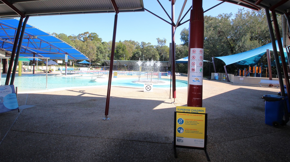 Inside the building, there is a narrow breezeway that serves as a control point for guests entering and exiting. A black pool fence acts as a barrier, indicating that entry requires payment. To the right, there is a low reception counter with a small selection of retail items, such as swimming costumes and goggles. Attached to the reception area is a cozy little café that extends towards the right. Inside the building, there is a narrow breezeway that serves as a control point for guests entering and exiting. A black pool fence acts as a barrier, indicating that entry requires payment. To the right, there is a low reception counter with a small selection of retail items, such as swimming costumes and goggles. Attached to the reception area is a cozy little café that extends towards the right.