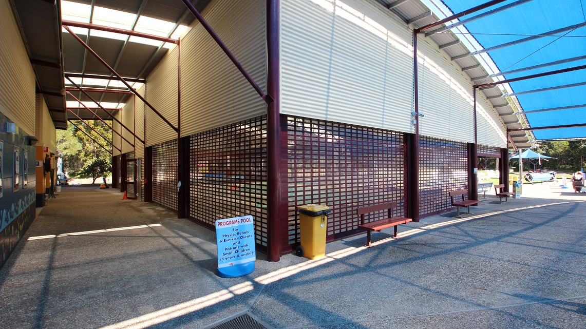 The back angle of the indoor heated pool. Three wooden chairs were placed just outside the pool. There's also a yellow rubbish bin nearby, as well as signage that provides information about pool programs. From this angle, the curved, corrugated iron roof lines are easily noticeable, giving the area a distinct appearance. The back corner of the pool area looks well-kept and tidy. The back angle of the indoor heated pool. Three wooden chairs were placed just outside the pool. There's also a yellow rubbish bin nearby, as well as signage that provides information about pool programs. From this angle, the curved, corrugated iron roof lines are easily noticeable, giving the area a distinct appearance. The back corner of the pool area looks well-kept and tidy.