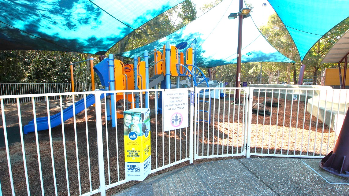 The playground area of the open-air aquatic pool. It's surrounded by trees and covered by white fences. There are signs reminding visitors to keep a close eye on their children. To provide some shade from the sun, three wide pieces of cloth are placed over the area. In the center, there is a post with spotlights, adding some illumination to the space. The playground area of the open-air aquatic pool. It's surrounded by trees and covered by white fences. There are signs reminding visitors to keep a close eye on their children. To provide some shade from the sun, three wide pieces of cloth are placed over the area. In the center, there is a post with spotlights, adding some illumination to the space.
