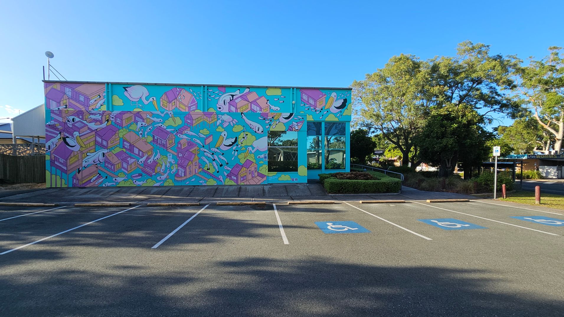 A vibrant mural covers the side of a building, featuring colourful houses and abstract shapes on a bright blue background. To the right, there are large windows reflecting the surrounding greenery. In the foreground, a parking area with marked spaces, including accessible parking spots, is visible. Trees and bushes are in the background, providing a natural setting around the building. A vibrant mural covers the side of a building, featuring colourful houses and abstract shapes on a bright blue background. To the right, there are large windows reflecting the surrounding greenery. In the foreground, a parking area with marked spaces, including accessible parking spots, is visible. Trees and bushes are in the background, providing a natural setting around the building.