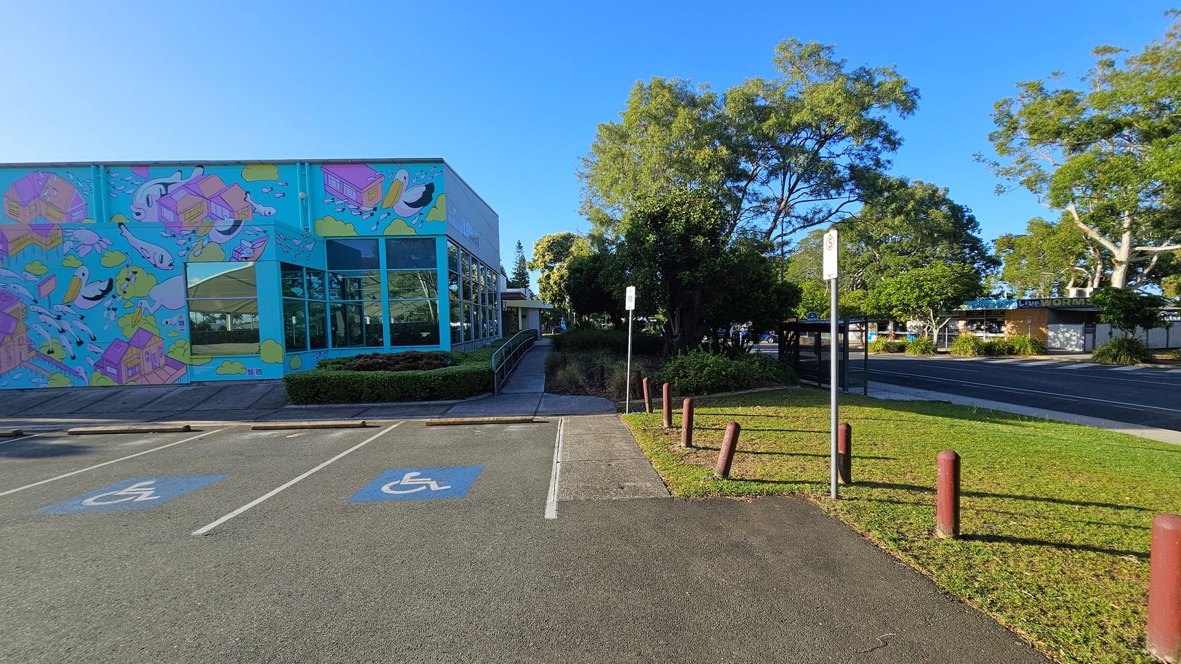 A colourful building on the left features a mural with whimsical designs, including fish and houses. Large windows reflect the surrounding greenery. In front, there are two marked disabled parking spaces. A pathway leads from the parking area towards a grassy section with bollards and a signpost. On the right, trees provide shade, and a road runs parallel to the building. A bus stop and more buildings are visible in the background. A colourful building on the left features a mural with whimsical designs, including fish and houses. Large windows reflect the surrounding greenery. In front, there are two marked disabled parking spaces. A pathway leads from the parking area towards a grassy section with bollards and a signpost. On the right, trees provide shade, and a road runs parallel to the building. A bus stop and more buildings are visible in the background.