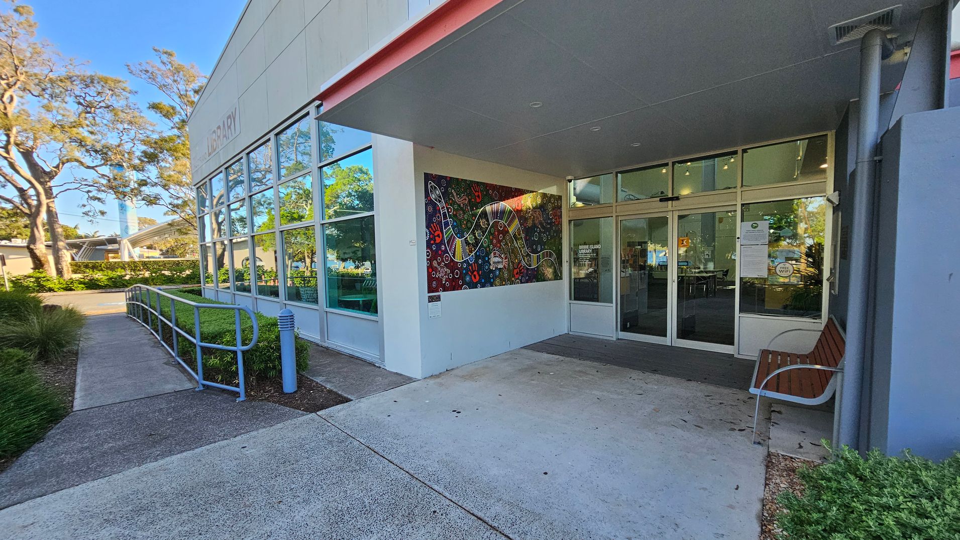 The image shows the entrance to a building with large glass windows and a colourful mural on the wall to the right. A bench is positioned near the entrance, and a pathway with a railing leads up to the door. The building is surrounded by greenery, with trees visible in the background. The ceiling above the entrance is grey, and the ground is paved with concrete. The image shows the entrance to a building with large glass windows and a colourful mural on the wall to the right. A bench is positioned near the entrance, and a pathway with a railing leads up to the door. The building is surrounded by greenery, with trees visible in the background. The ceiling above the entrance is grey, and the ground is paved with concrete.