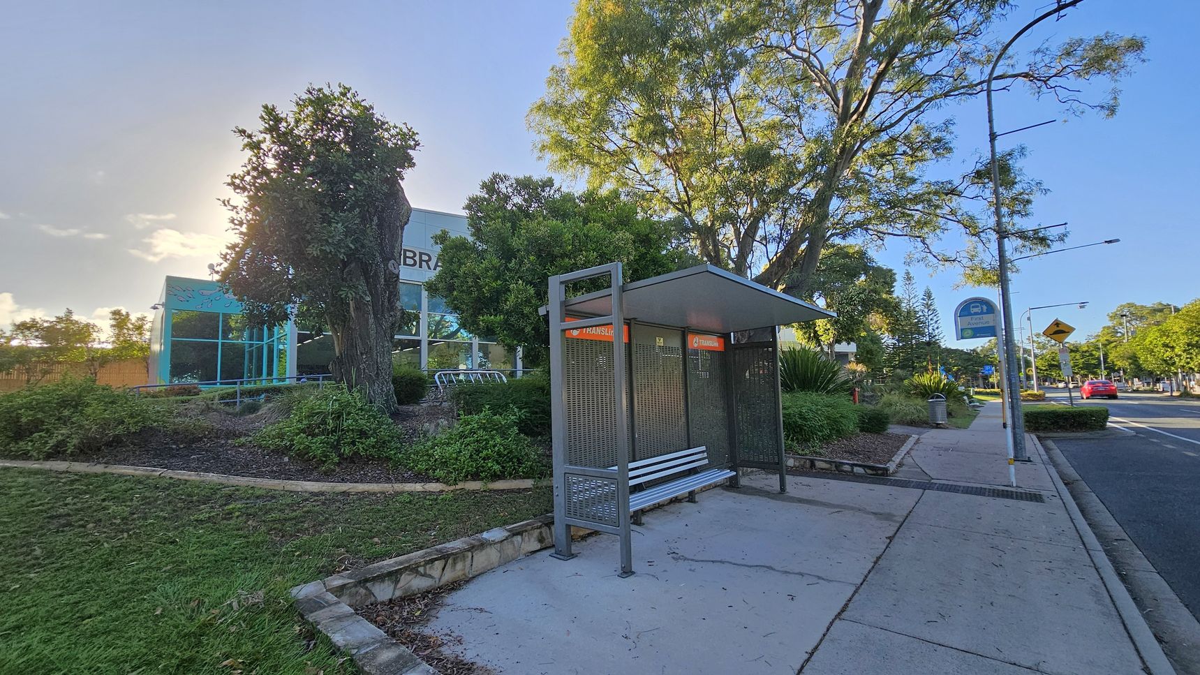 A bus stop with a metal bench and shelter is on the right, next to a paved footpath. A street sign and a rubbish bin are nearby. On the left, a building with large glass windows and the word "LIBRARY" is partially visible, surrounded by trees and landscaped greenery. A large tree stands prominently in the centre. The road runs parallel to the footpath, with a red car in the distance. The sky is clear, and the area is well-lit. A bus stop with a metal bench and shelter is on the right, next to a paved footpath. A street sign and a rubbish bin are nearby. On the left, a building with large glass windows and the word "LIBRARY" is partially visible, surrounded by trees and landscaped greenery. A large tree stands prominently in the centre. The road runs parallel to the footpath, with a red car in the distance. The sky is clear, and the area is well-lit.