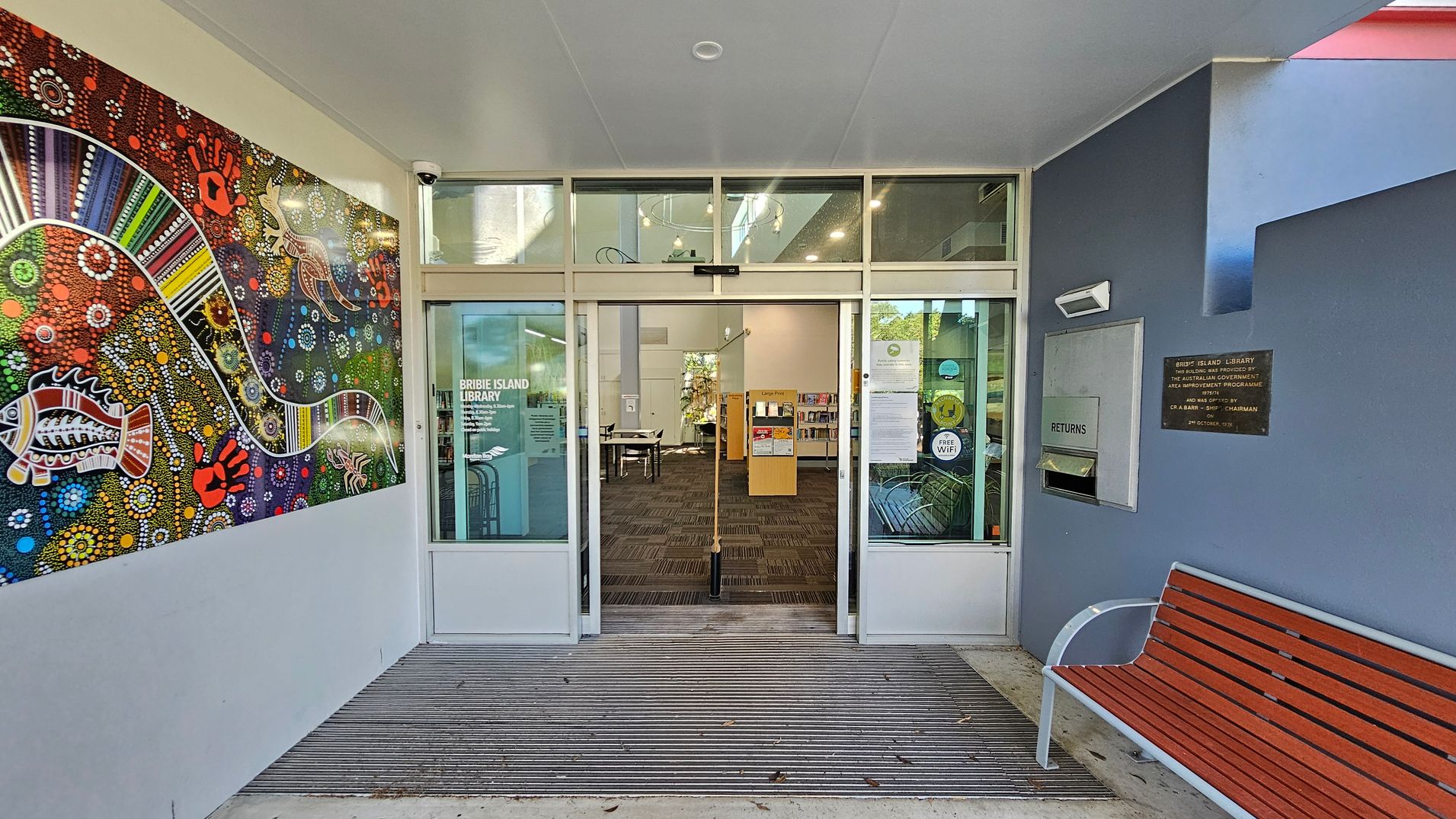 A library entrance with glass doors in the centre, labelled "Bribie Island Library". To the left, a vibrant mural features colourful Aboriginal art with patterns and animal motifs. On the right, a blue wall has a book return slot and a plaque. A wooden bench with metal armrests is positioned below. The floor is a combination of concrete and a ribbed mat leading into the library. The ceiling is plain with a light fixture. A library entrance with glass doors in the centre, labelled "Bribie Island Library". To the left, a vibrant mural features colourful Aboriginal art with patterns and animal motifs. On the right, a blue wall has a book return slot and a plaque. A wooden bench with metal armrests is positioned below. The floor is a combination of concrete and a ribbed mat leading into the library. The ceiling is plain with a light fixture.