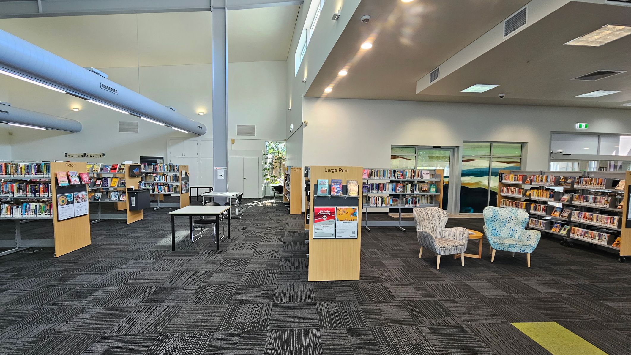 A library interior with a grey carpeted floor. On the left, there are bookshelves labelled "Fiction" filled with books. In the centre, a section labelled "Large Print" is visible, along with a couple of tables and chairs. On the right, there are more bookshelves and two patterned armchairs with a small round table between them. The ceiling has a mix of recessed and fluorescent lighting, and large windows allow natural light in. A library interior with a grey carpeted floor. On the left, there are bookshelves labelled "Fiction" filled with books. In the centre, a section labelled "Large Print" is visible, along with a couple of tables and chairs. On the right, there are more bookshelves and two patterned armchairs with a small round table between them. The ceiling has a mix of recessed and fluorescent lighting, and large windows allow natural light in.