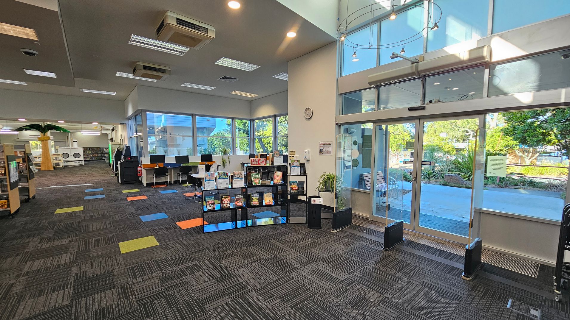 A library interior with a patterned carpet featuring colourful squares. On the right, large glass doors lead outside, with greenery visible through the windows. In the centre, a display shelf holds various books. To the left, a row of computers is set up on desks, and further back, bookshelves and a decorative palm tree are visible. The ceiling has recessed lighting and air conditioning units. A library interior with a patterned carpet featuring colourful squares. On the right, large glass doors lead outside, with greenery visible through the windows. In the centre, a display shelf holds various books. To the left, a row of computers is set up on desks, and further back, bookshelves and a decorative palm tree are visible. The ceiling has recessed lighting and air conditioning units.