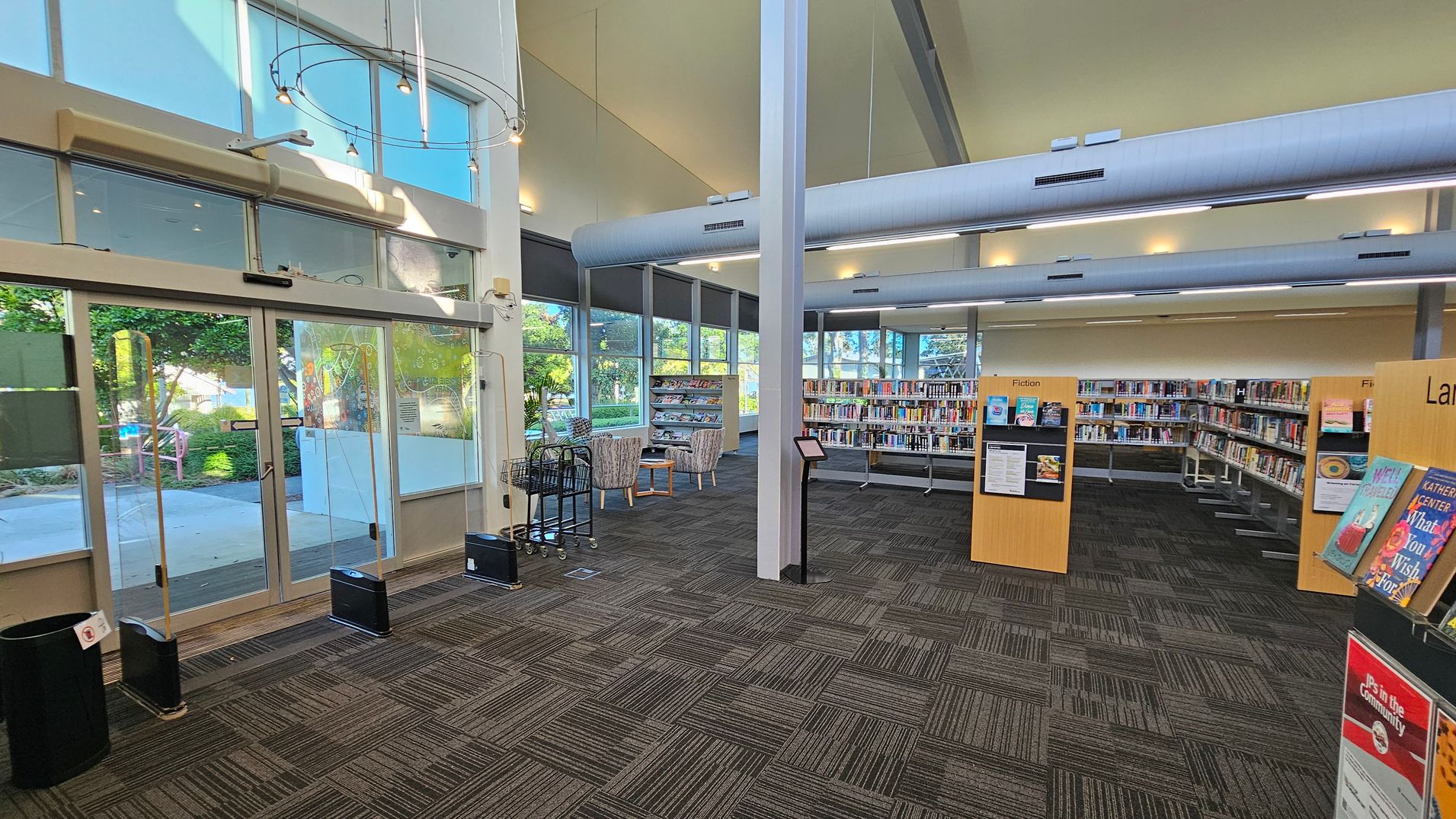 A library interior with large glass doors on the left, leading outside. The floor is covered with patterned carpet tiles in shades of grey and green. In the centre, there are bookshelves filled with books, labelled "Fiction." To the right, more bookshelves and a display stand with books. The ceiling is high with exposed ducts and modern lighting. Large windows on the far wall let in natural light, and there are two armchairs with a small table near the windows. A library interior with large glass doors on the left, leading outside. The floor is covered with patterned carpet tiles in shades of grey and green. In the centre, there are bookshelves filled with books, labelled "Fiction." To the right, more bookshelves and a display stand with books. The ceiling is high with exposed ducts and modern lighting. Large windows on the far wall let in natural light, and there are two armchairs with a small table near the windows.