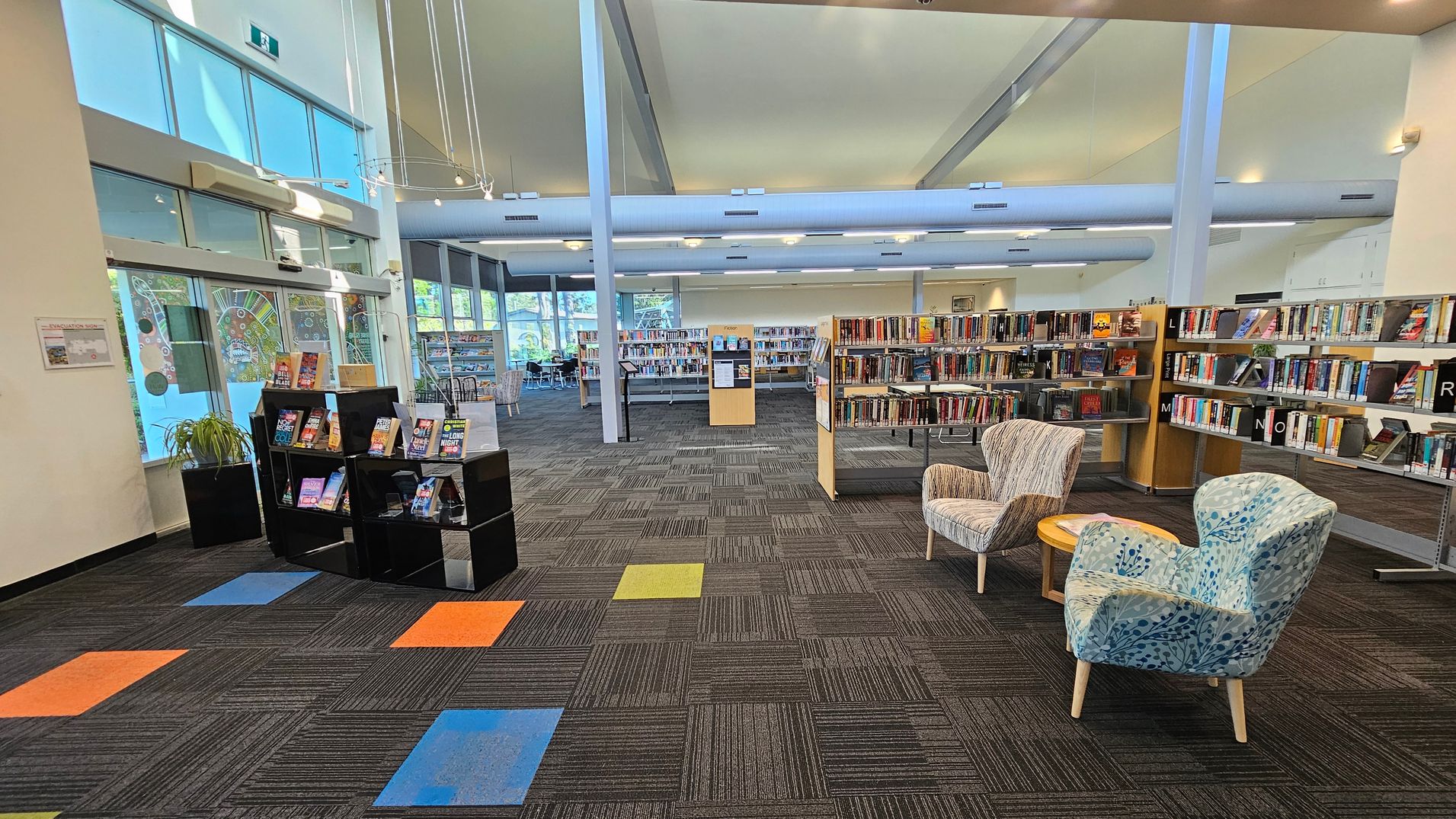 A library interior with a modern design. On the left, there are large windows with decorative patterns and a black display shelf holding books. The floor has a patterned carpet with colourful square accents in orange, blue, and green. In the centre, there are bookshelves filled with books. On the right, two patterned armchairs and a small round table are placed near more bookshelves. The ceiling is high with exposed beams and hanging lights. A library interior with a modern design. On the left, there are large windows with decorative patterns and a black display shelf holding books. The floor has a patterned carpet with colourful square accents in orange, blue, and green. In the centre, there are bookshelves filled with books. On the right, two patterned armchairs and a small round table are placed near more bookshelves. The ceiling is high with exposed beams and hanging lights.