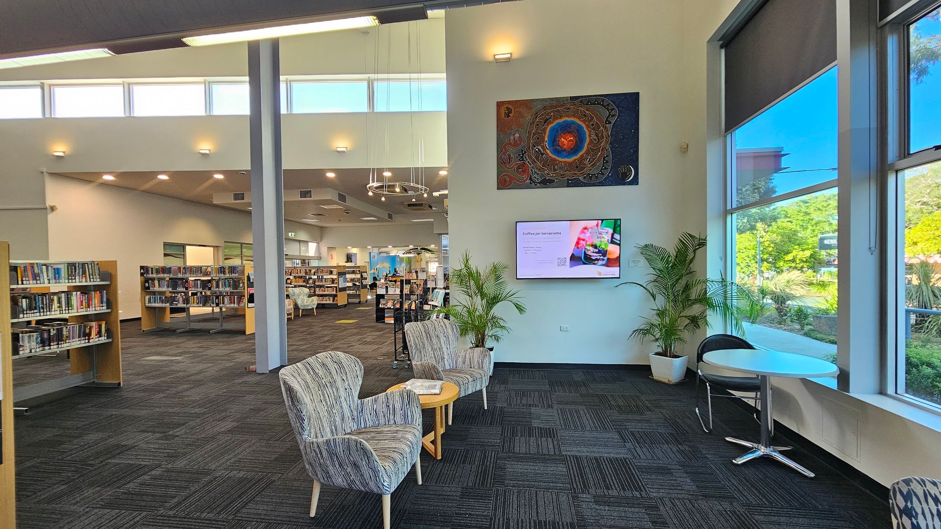 A modern library interior with a spacious layout. On the left, there are bookshelves filled with books. In the centre, two patterned armchairs and a small round table are placed near a wall with a digital screen and artwork above it. On the right, large windows allow natural light to illuminate the space, with a small round table and chair nearby. The floor is carpeted in a dark pattern, and the ceiling features recessed lighting and a circular chandelier. A modern library interior with a spacious layout. On the left, there are bookshelves filled with books. In the centre, two patterned armchairs and a small round table are placed near a wall with a digital screen and artwork above it. On the right, large windows allow natural light to illuminate the space, with a small round table and chair nearby. The floor is carpeted in a dark pattern, and the ceiling features recessed lighting and a circular chandelier.