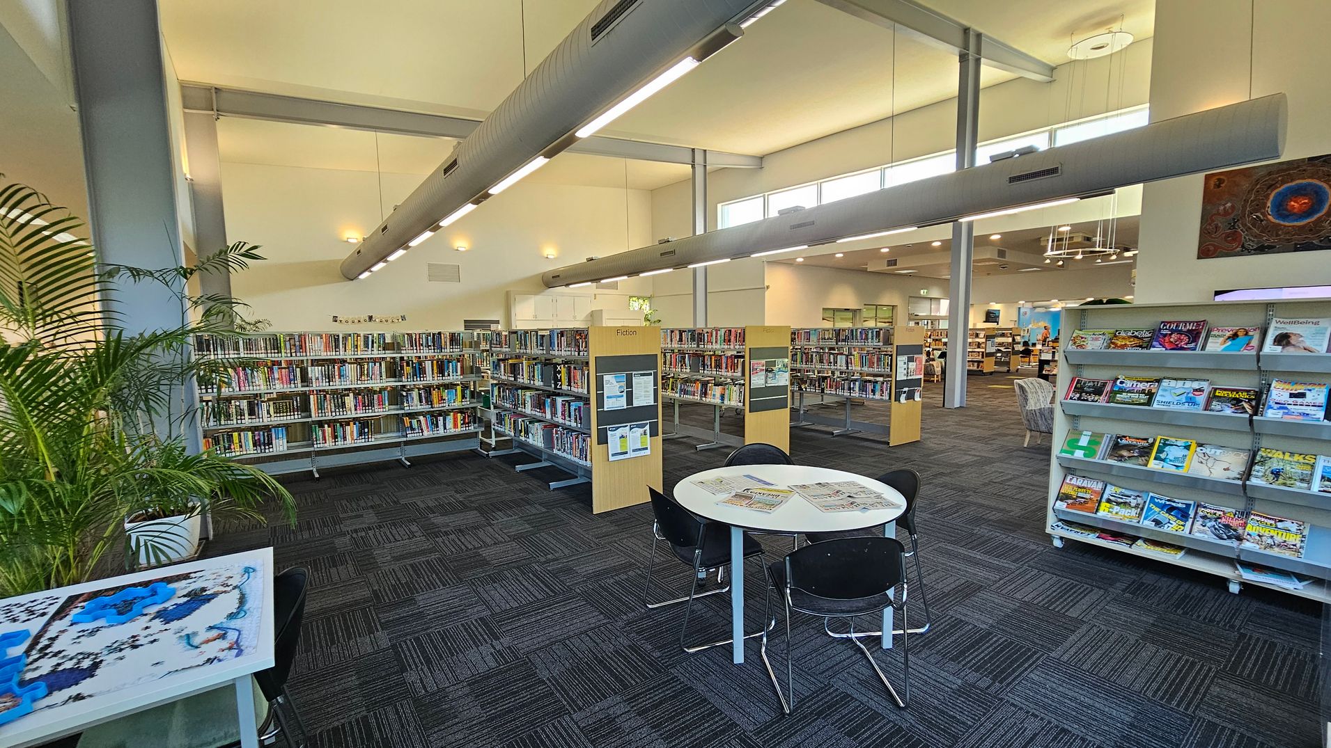 A spacious library interior with a high ceiling and large windows. On the left, there are tall bookshelves filled with books and a potted plant. In the centre, a round table with chairs has newspapers on it. On the right, a magazine rack displays various colourful magazines. The floor is carpeted with a dark pattern, and overhead, long light fixtures hang from the ceiling. In the background, more bookshelves and seating areas are visible. A spacious library interior with a high ceiling and large windows. On the left, there are tall bookshelves filled with books and a potted plant. In the centre, a round table with chairs has newspapers on it. On the right, a magazine rack displays various colourful magazines. The floor is carpeted with a dark pattern, and overhead, long light fixtures hang from the ceiling. In the background, more bookshelves and seating areas are visible.