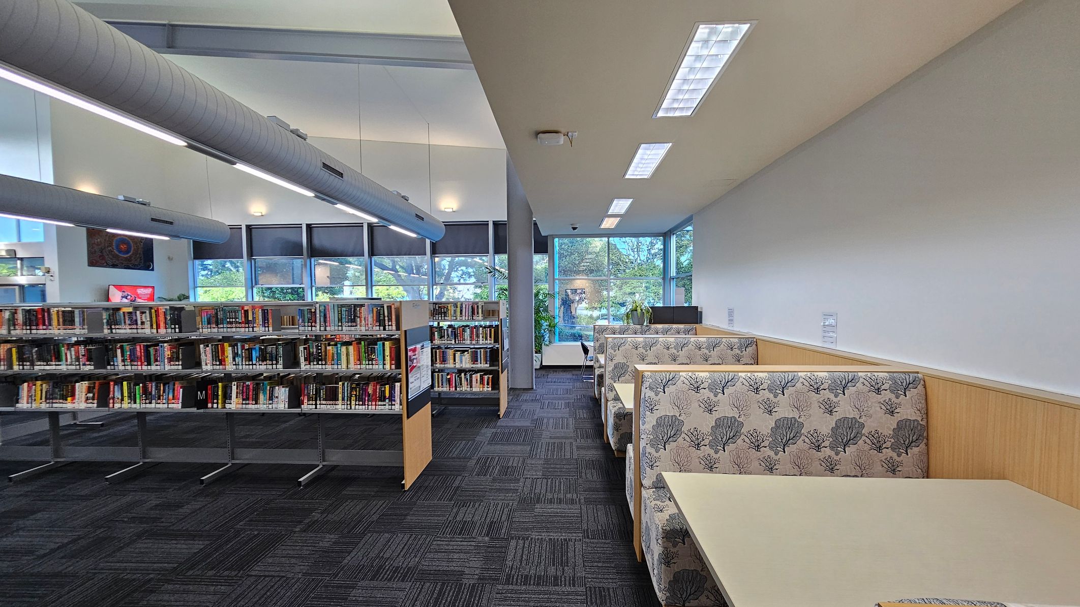 Library interior with bookshelves on the left filled with books, and a row of cushioned booths on the right with floral-patterned upholstery. The floor is carpeted in a dark, patterned design. Large windows in the background allow natural light to enter, with views of trees outside. The ceiling features long, cylindrical air ducts and recessed lighting. Library interior with bookshelves on the left filled with books, and a row of cushioned booths on the right with floral-patterned upholstery. The floor is carpeted in a dark, patterned design. Large windows in the background allow natural light to enter, with views of trees outside. The ceiling features long, cylindrical air ducts and recessed lighting.