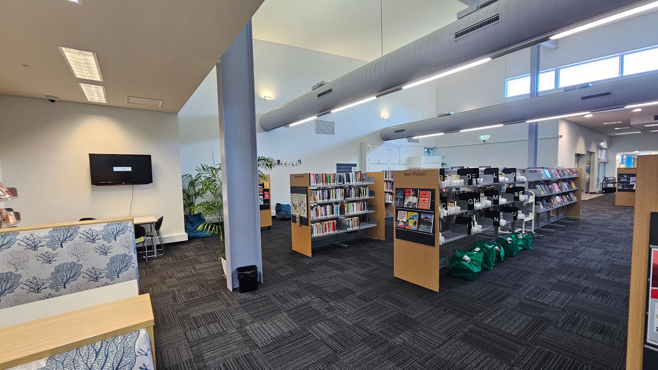 A library interior with a dark carpeted floor. On the left, a wall-mounted TV above a small table and chairs, with a decorative panel featuring blue coral patterns. In the centre, bookshelves labelled "Biography" and "Non Fiction" are filled with books. On the right, more shelves with alphabetic dividers and green bags on the floor. The ceiling has large cylindrical ducts and fluorescent lights. Tall windows on the right allow natural light in. A library interior with a dark carpeted floor. On the left, a wall-mounted TV above a small table and chairs, with a decorative panel featuring blue coral patterns. In the centre, bookshelves labelled "Biography" and "Non Fiction" are filled with books. On the right, more shelves with alphabetic dividers and green bags on the floor. The ceiling has large cylindrical ducts and fluorescent lights. Tall windows on the right allow natural light in.