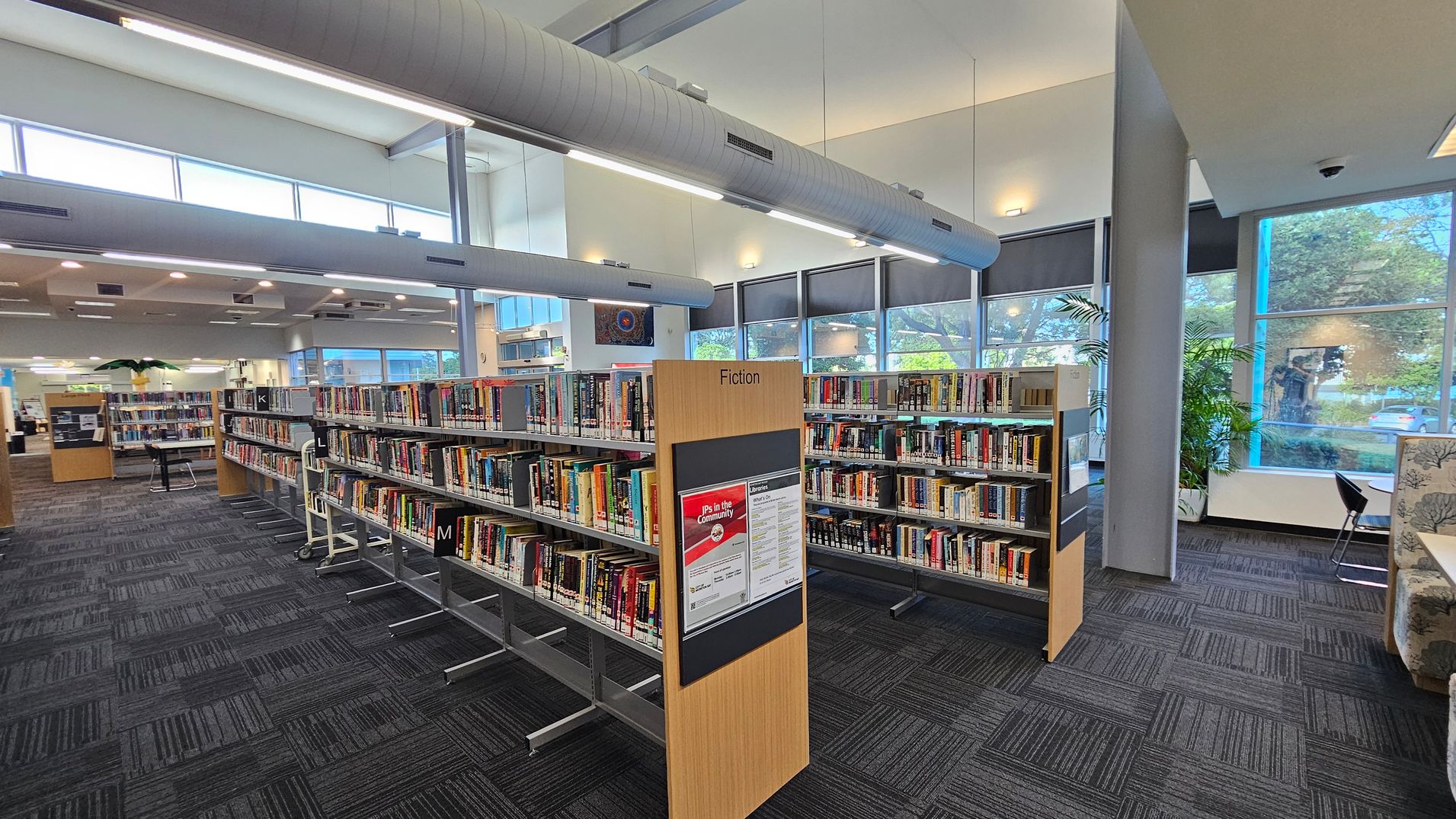 Bookshelves filled with books are arranged in rows on a carpeted floor, with a sign labelled "Fiction" in the foreground. Large windows on the right allow natural light to illuminate the space, and there are potted plants near the windows. The ceiling features exposed ductwork and fluorescent lighting. In the background, more bookshelves and a seating area with chairs and tables are visible. Bookshelves filled with books are arranged in rows on a carpeted floor, with a sign labelled "Fiction" in the foreground. Large windows on the right allow natural light to illuminate the space, and there are potted plants near the windows. The ceiling features exposed ductwork and fluorescent lighting. In the background, more bookshelves and a seating area with chairs and tables are visible.