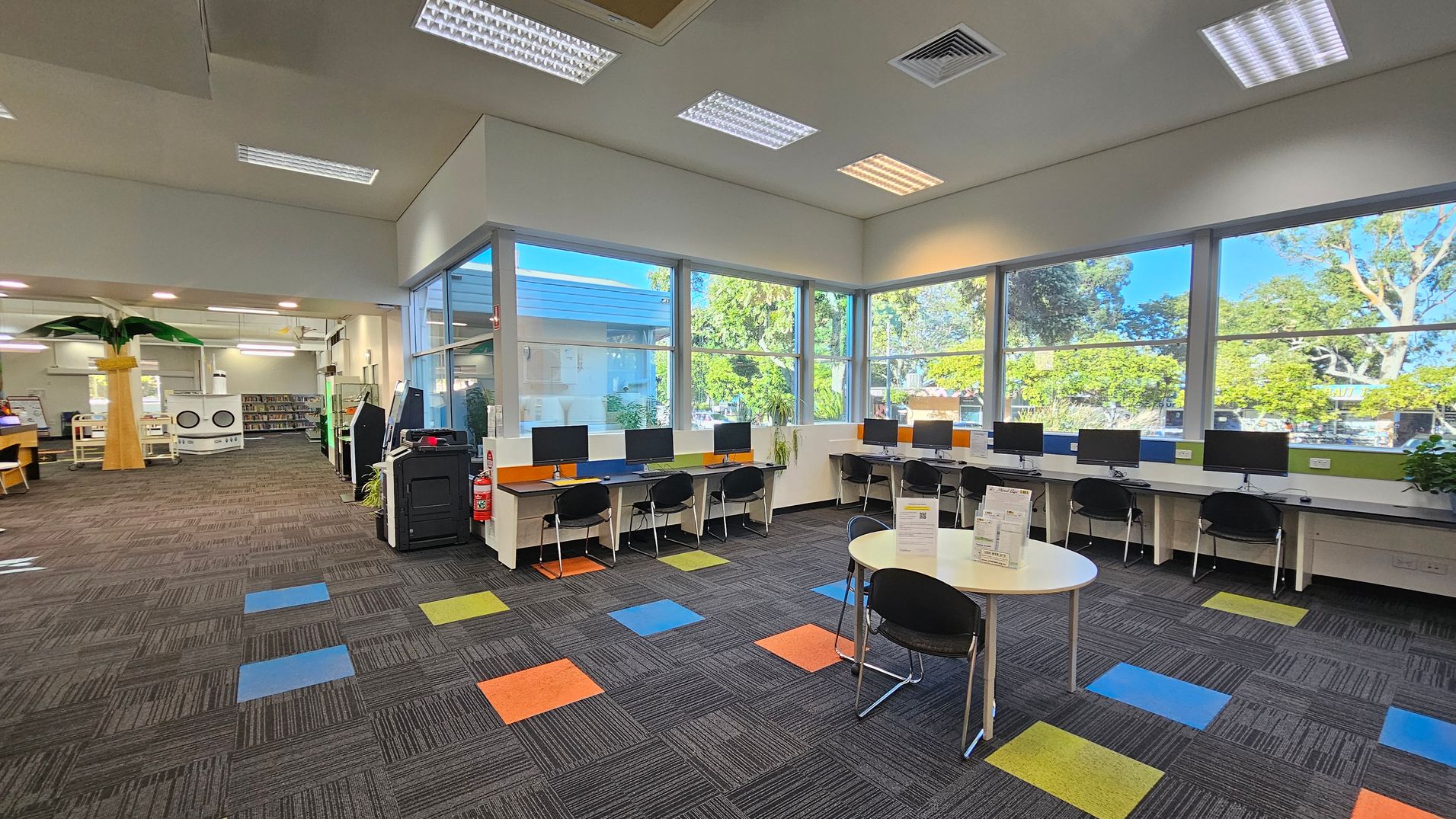 A modern library interior with large windows on the right, allowing natural light to illuminate the space. Several computer workstations with black chairs are lined up against the windows. In the foreground, a round table with informational pamphlets and chairs is visible. The floor is carpeted with a pattern of grey tiles interspersed with colourful squares. On the left, there is a tall artificial palm tree and bookshelves. The ceiling has recessed lighting and air vents. A modern library interior with large windows on the right, allowing natural light to illuminate the space. Several computer workstations with black chairs are lined up against the windows. In the foreground, a round table with informational pamphlets and chairs is visible. The floor is carpeted with a pattern of grey tiles interspersed with colourful squares. On the left, there is a tall artificial palm tree and bookshelves. The ceiling has recessed lighting and air vents.