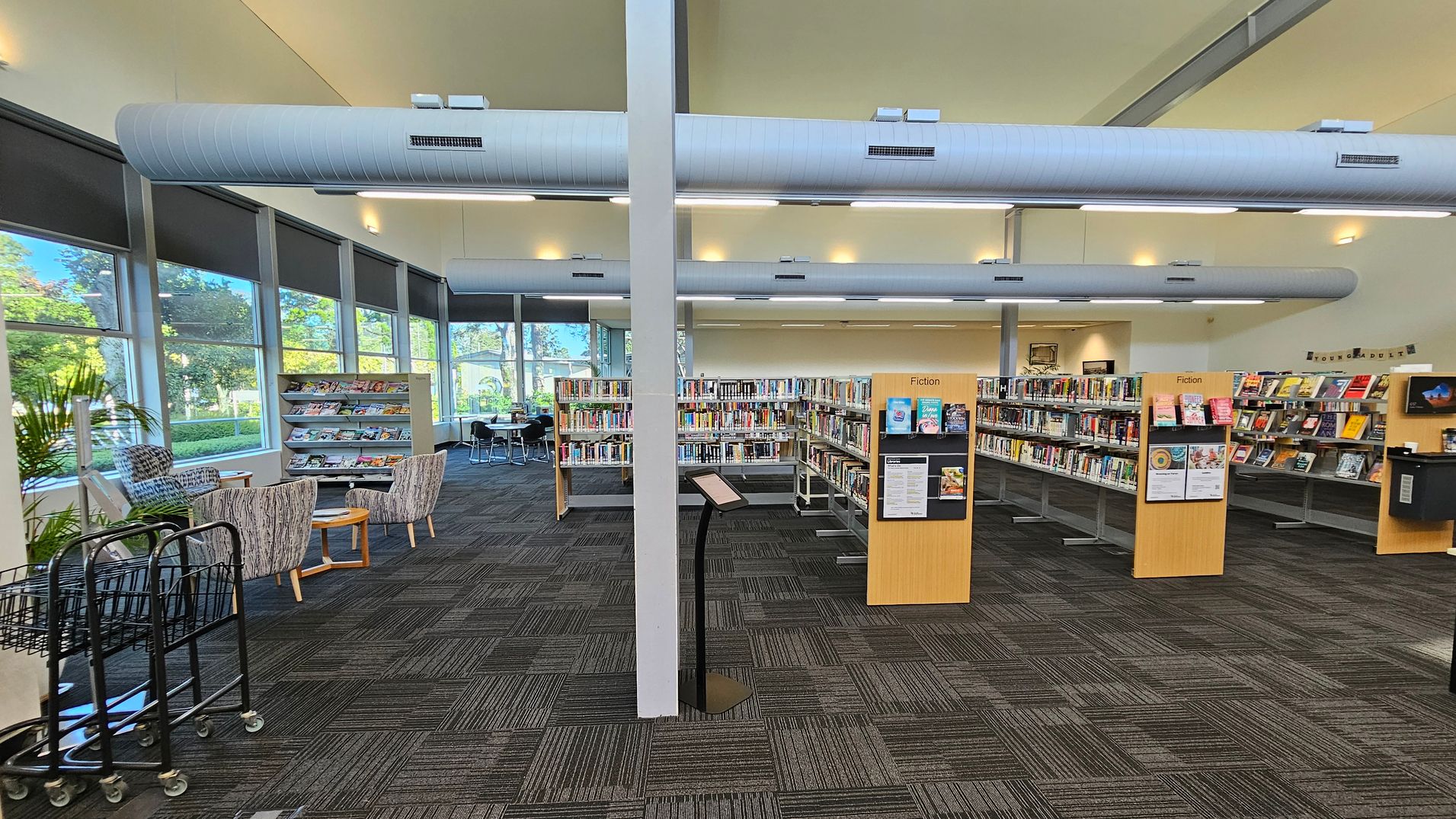 A library interior with a grey and green patterned carpet. On the left, there are large windows with roller blinds, a magazine rack, and seating with patterned chairs and a small table. In the centre, a vertical support beam stands in front of a digital kiosk. Bookshelves labelled "Fiction" are filled with books. On the right, more bookshelves and a section labelled "Young Adult" are visible. The ceiling features large cylindrical air ducts and recessed lighting. A library interior with a grey and green patterned carpet. On the left, there are large windows with roller blinds, a magazine rack, and seating with patterned chairs and a small table. In the centre, a vertical support beam stands in front of a digital kiosk. Bookshelves labelled "Fiction" are filled with books. On the right, more bookshelves and a section labelled "Young Adult" are visible. The ceiling features large cylindrical air ducts and recessed lighting.
