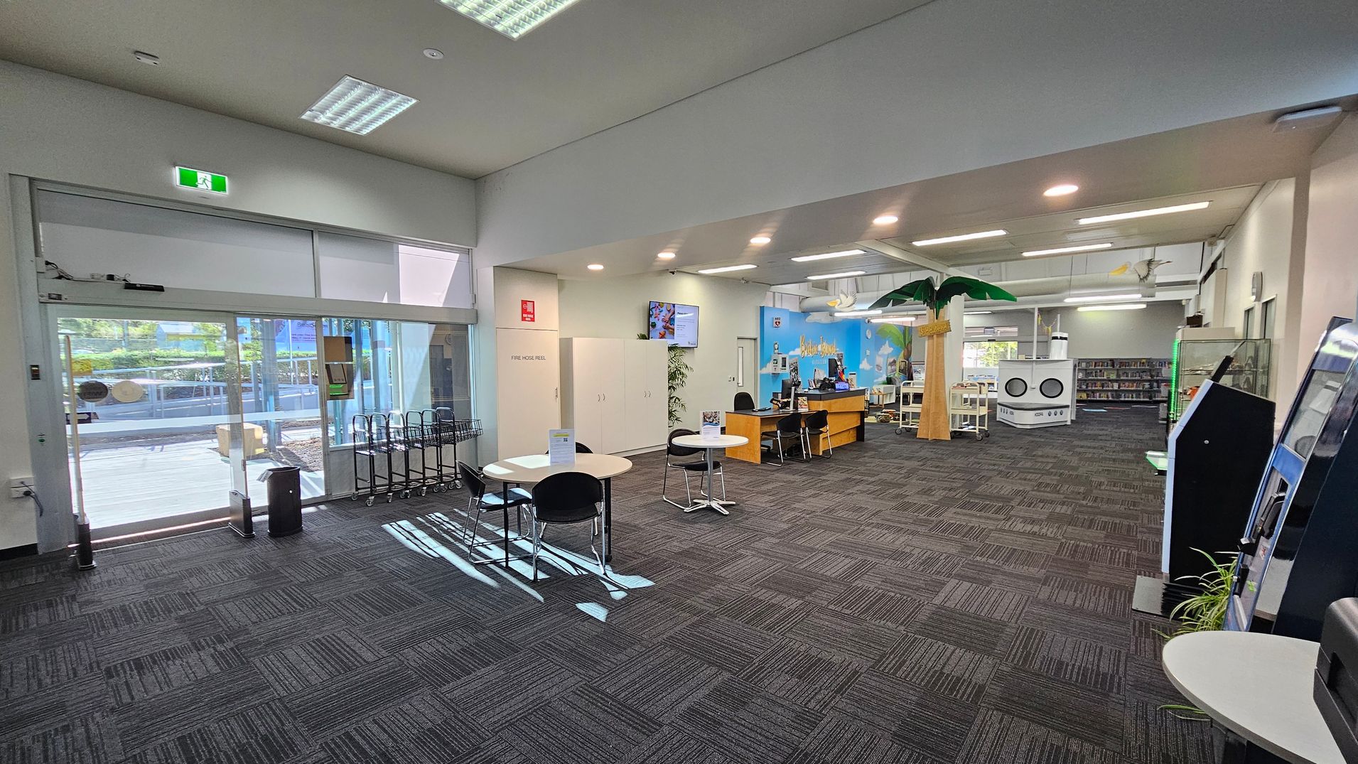 A spacious interior with a grey carpeted floor. On the left, there are large glass doors leading outside, with a row of trolleys nearby. In the centre, there are round tables with chairs. Towards the back, a reception desk is visible, with a colourful mural on the wall behind it. On the right, there are self-service machines and a display case. The ceiling is equipped with fluorescent lights, and a decorative palm tree stands near the centre. Bookshelves are visible in the background. A spacious interior with a grey carpeted floor. On the left, there are large glass doors leading outside, with a row of trolleys nearby. In the centre, there are round tables with chairs. Towards the back, a reception desk is visible, with a colourful mural on the wall behind it. On the right, there are self-service machines and a display case. The ceiling is equipped with fluorescent lights, and a decorative palm tree stands near the centre. Bookshelves are visible in the background.