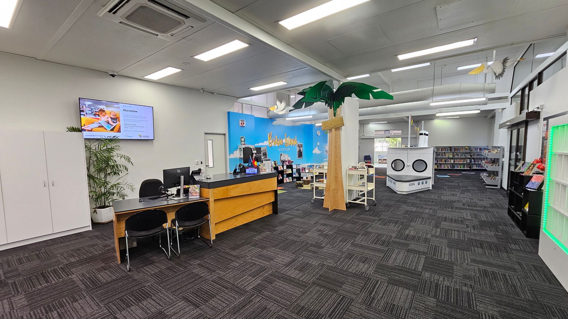 A library interior with a grey carpeted floor and a white ceiling with fluorescent lights. On the left, there's a reception desk with computers and chairs, and a potted plant nearby. A wall-mounted screen displays information. In the centre, a large decorative palm tree stands next to a blue wall with "Bribie Island" written on it. Bookshelves and a white cylindrical structure resembling a ship are visible in the background. On the right, more bookshelves and a display with green lighting. A library interior with a grey carpeted floor and a white ceiling with fluorescent lights. On the left, there's a reception desk with computers and chairs, and a potted plant nearby. A wall-mounted screen displays information. In the centre, a large decorative palm tree stands next to a blue wall with "Bribie Island" written on it. Bookshelves and a white cylindrical structure resembling a ship are visible in the background. On the right, more bookshelves and a display with green lighting.