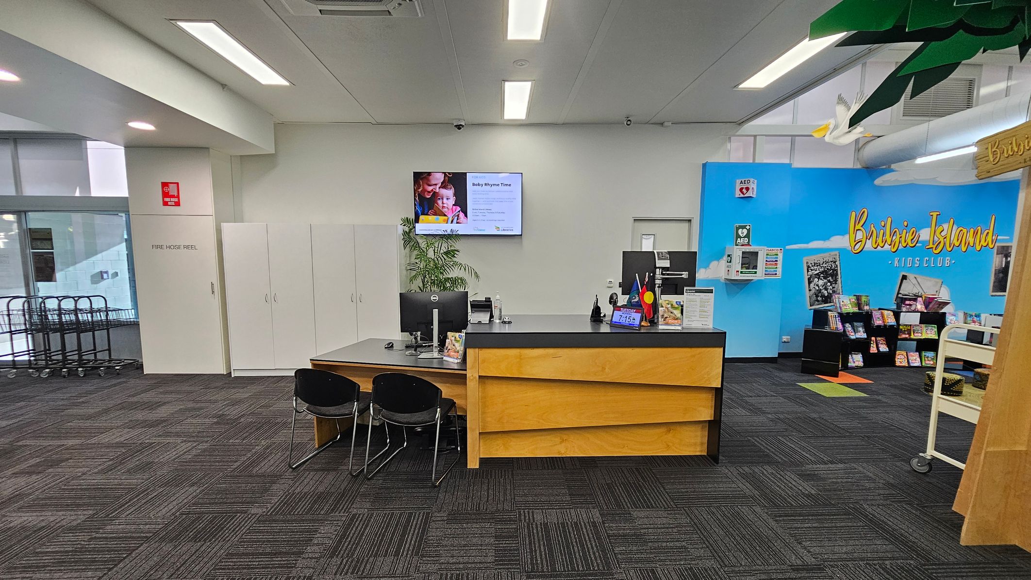 A library interior with a reception desk in the centre, featuring two black chairs and computer monitors. Behind the desk, a wall-mounted screen displays information. To the left, a fire hose reel is marked on a white door, with trolleys near a window. On the right, a blue wall with "Bribie Island Kids Club" text and a display of books. The ceiling has fluorescent lights, and the floor is covered with dark patterned carpet tiles. A white bird decoration hangs from the ceiling. A library interior with a reception desk in the centre, featuring two black chairs and computer monitors. Behind the desk, a wall-mounted screen displays information. To the left, a fire hose reel is marked on a white door, with trolleys near a window. On the right, a blue wall with "Bribie Island Kids Club" text and a display of books. The ceiling has fluorescent lights, and the floor is covered with dark patterned carpet tiles. A white bird decoration hangs from the ceiling.