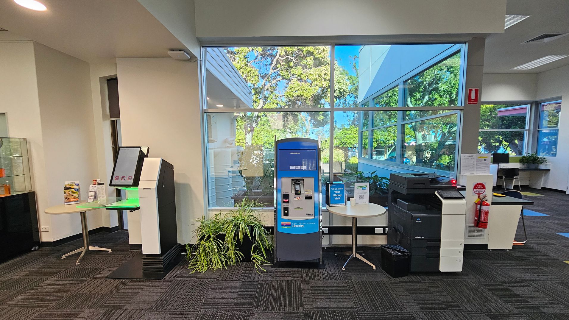 A room with a large window in the background showing trees outside. On the left, there's a kiosk with a screen and a small round table with brochures. In the centre, a blue and grey machine is flanked by potted plants. On the right, there's a printer with a fire extinguisher nearby. The floor is carpeted with a dark pattern, and the ceiling has recessed lighting. A room with a large window in the background showing trees outside. On the left, there's a kiosk with a screen and a small round table with brochures. In the centre, a blue and grey machine is flanked by potted plants. On the right, there's a printer with a fire extinguisher nearby. The floor is carpeted with a dark pattern, and the ceiling has recessed lighting.