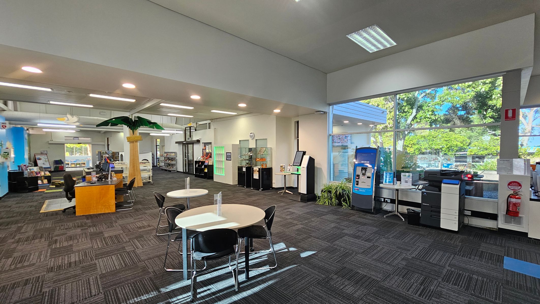 A spacious interior with a high ceiling and large windows on the right, allowing natural light to illuminate the room. In the foreground, there are round tables with black chairs. To the right, a blue kiosk and a printer are near a fire extinguisher. The left side features a reception desk with chairs and a decorative palm tree. Bookshelves and display cases line the walls, and the floor is covered with patterned carpet tiles. A spacious interior with a high ceiling and large windows on the right, allowing natural light to illuminate the room. In the foreground, there are round tables with black chairs. To the right, a blue kiosk and a printer are near a fire extinguisher. The left side features a reception desk with chairs and a decorative palm tree. Bookshelves and display cases line the walls, and the floor is covered with patterned carpet tiles.