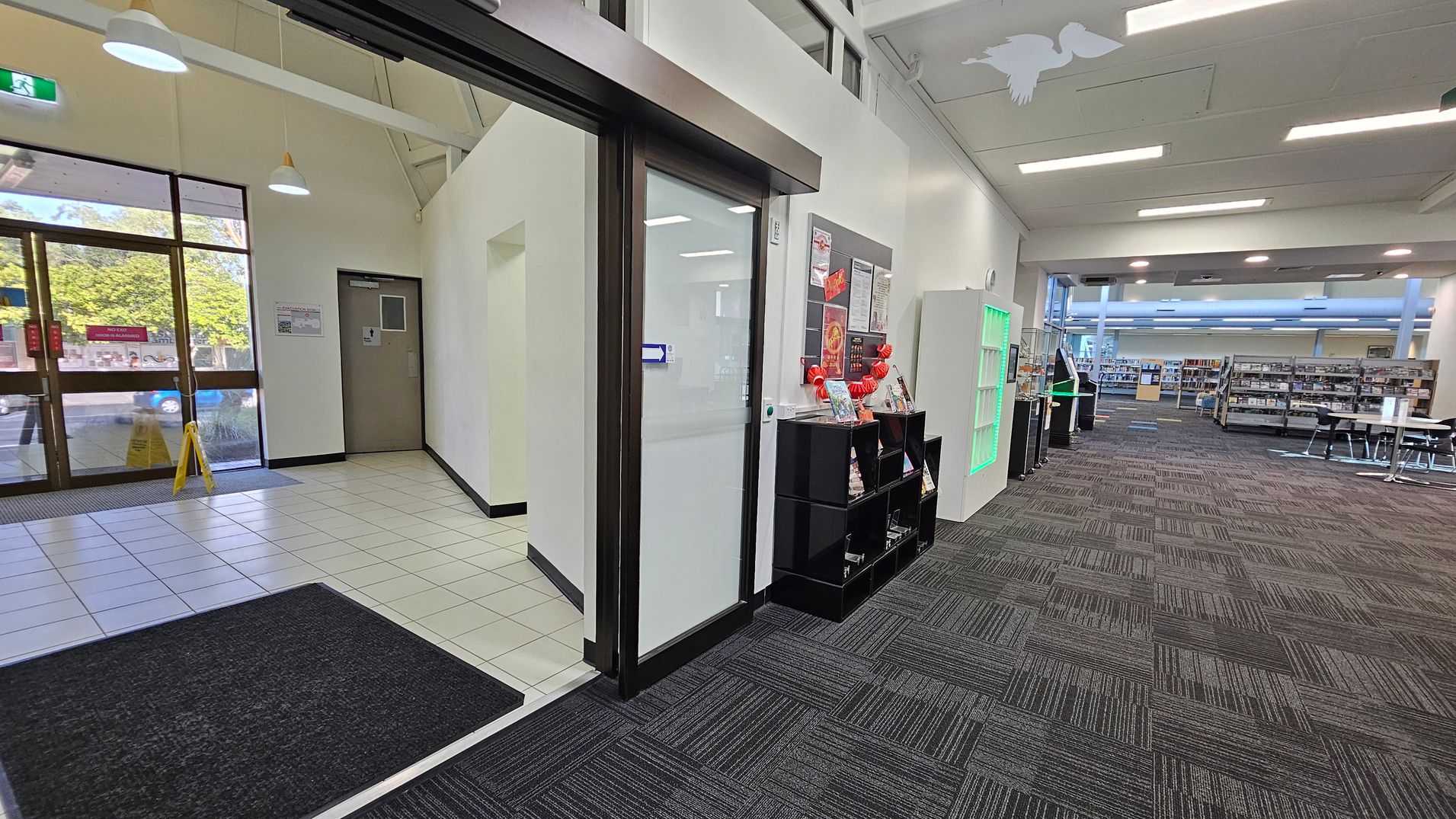 On the left, there is a glass entrance door with a tiled floor and a caution sign. A door is visible on the adjacent wall. In the centre, a glass door leads into a carpeted area with a black shelving unit displaying brochures and a bulletin board above. On the right, the space opens into a library with bookshelves, tables, and chairs. The ceiling has fluorescent lights, and a green-lit display case is near the entrance. On the left, there is a glass entrance door with a tiled floor and a caution sign. A door is visible on the adjacent wall. In the centre, a glass door leads into a carpeted area with a black shelving unit displaying brochures and a bulletin board above. On the right, the space opens into a library with bookshelves, tables, and chairs. The ceiling has fluorescent lights, and a green-lit display case is near the entrance.