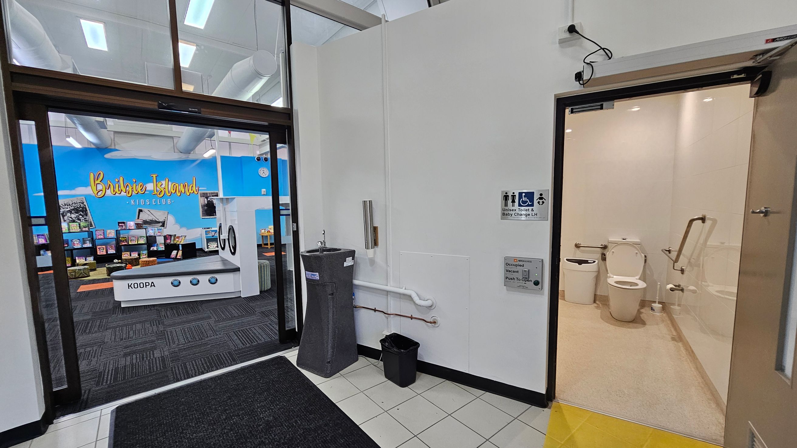 A hallway with a black mat on a tiled floor. On the left, there's an entrance to a room with a blue wall displaying "Bribie Island Kids Club" and shelves filled with books. On the right, a door leads to a unisex toilet with a baby change facility, featuring a white toilet and handrails. A grey water fountain and a small black bin are mounted on the wall outside the toilet. The ceiling has visible ducts and lights. A hallway with a black mat on a tiled floor. On the left, there's an entrance to a room with a blue wall displaying "Bribie Island Kids Club" and shelves filled with books. On the right, a door leads to a unisex toilet with a baby change facility, featuring a white toilet and handrails. A grey water fountain and a small black bin are mounted on the wall outside the toilet. The ceiling has visible ducts and lights.