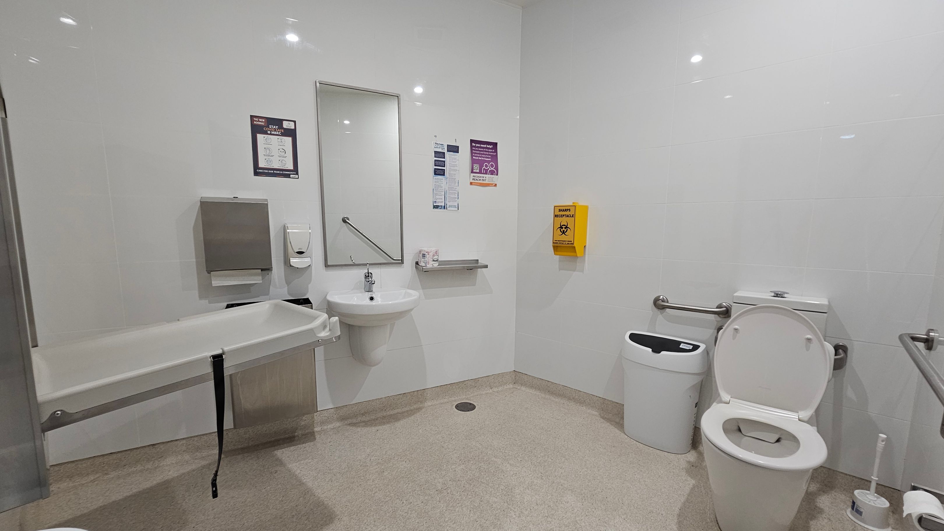 A clean, accessible bathroom with white walls and a light-coloured floor. On the left, a foldable changing table is mounted on the wall. In the centre, a sink with a mirror above it is flanked by a paper towel dispenser and a soap dispenser. A yellow sharps disposal box is mounted on the wall. On the right, a toilet with support rails is next to a bin and a toilet brush. The ceiling has recessed lighting. A clean, accessible bathroom with white walls and a light-coloured floor. On the left, a foldable changing table is mounted on the wall. In the centre, a sink with a mirror above it is flanked by a paper towel dispenser and a soap dispenser. A yellow sharps disposal box is mounted on the wall. On the right, a toilet with support rails is next to a bin and a toilet brush. The ceiling has recessed lighting.