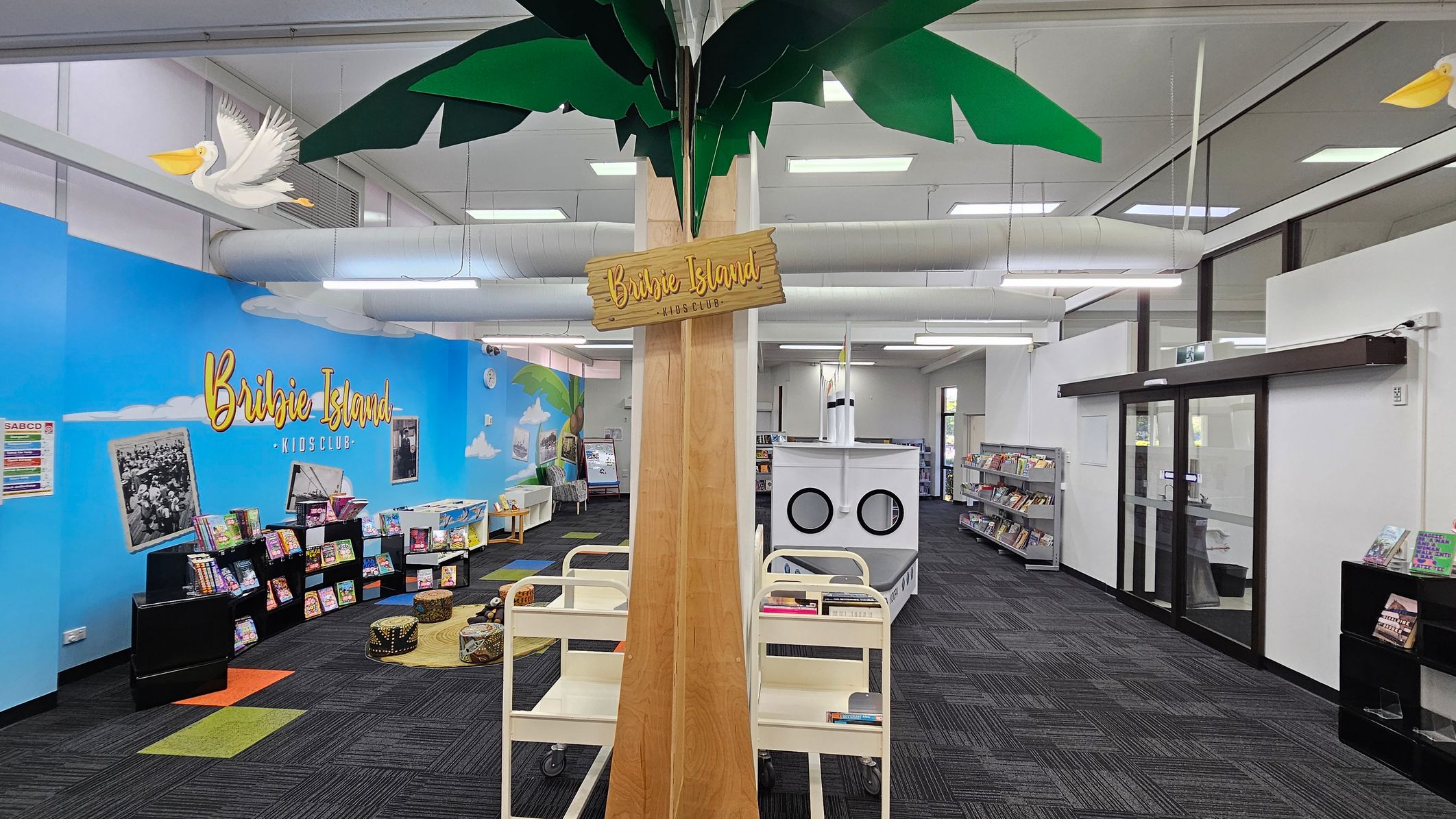 A library space with a central wooden column featuring a "Bribie Island Kids Club" sign. Above, green leaves and a pelican decoration are visible. On the left, a blue wall displays the same "Bribie Island" text, with books on black shelves and colourful seating on a patterned carpet. On the right, glass doors and more bookshelves are present. The ceiling has exposed ducts and fluorescent lights, and the floor is covered with dark carpet tiles. A library space with a central wooden column featuring a "Bribie Island Kids Club" sign. Above, green leaves and a pelican decoration are visible. On the left, a blue wall displays the same "Bribie Island" text, with books on black shelves and colourful seating on a patterned carpet. On the right, glass doors and more bookshelves are present. The ceiling has exposed ducts and fluorescent lights, and the floor is covered with dark carpet tiles.