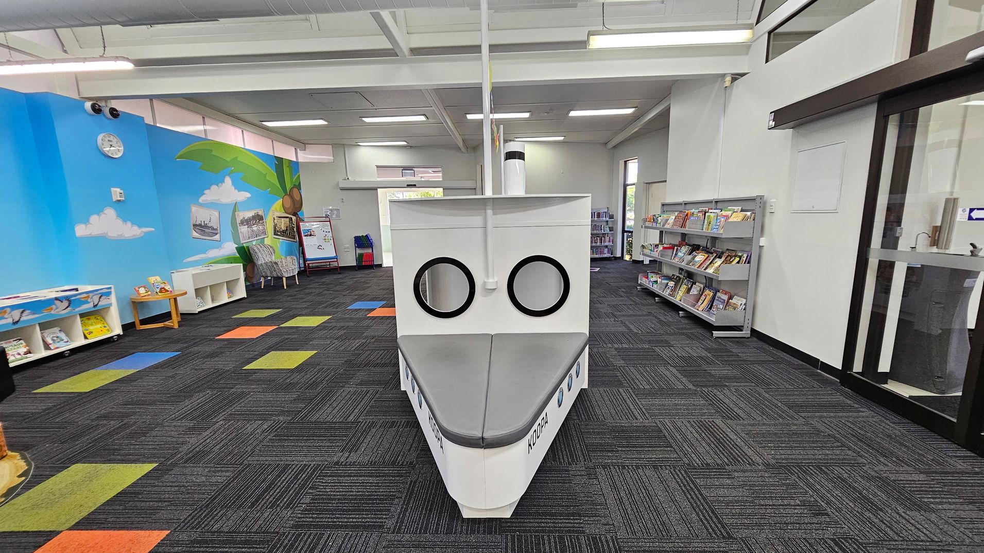 A library space with a boat-shaped reading nook in the centre, featuring two circular windows and grey seating. The left wall is painted with a blue sky, clouds, and a palm tree, with bookshelves and a clock. The right side has bookshelves filled with books and a glass door. The floor is carpeted in dark grey with colourful square patterns, and the ceiling has white panels with fluorescent lights. A library space with a boat-shaped reading nook in the centre, featuring two circular windows and grey seating. The left wall is painted with a blue sky, clouds, and a palm tree, with bookshelves and a clock. The right side has bookshelves filled with books and a glass door. The floor is carpeted in dark grey with colourful square patterns, and the ceiling has white panels with fluorescent lights.
