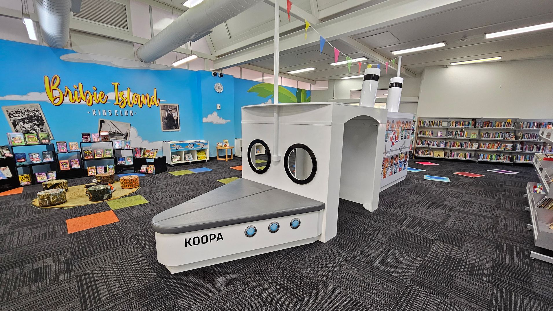 A children's library area with a nautical theme. On the left, a blue wall with "Bribie Island Kids Club" written in yellow, surrounded by bookshelves filled with colourful books. In the centre, a white play structure resembling a ship with portholes and labelled "KOOPA". The floor is carpeted in dark grey with colourful square accents. The ceiling has exposed ducts and fluorescent lighting. On the right, more bookshelves line the wall. A children's library area with a nautical theme. On the left, a blue wall with "Bribie Island Kids Club" written in yellow, surrounded by bookshelves filled with colourful books. In the centre, a white play structure resembling a ship with portholes and labelled "KOOPA". The floor is carpeted in dark grey with colourful square accents. The ceiling has exposed ducts and fluorescent lighting. On the right, more bookshelves line the wall.