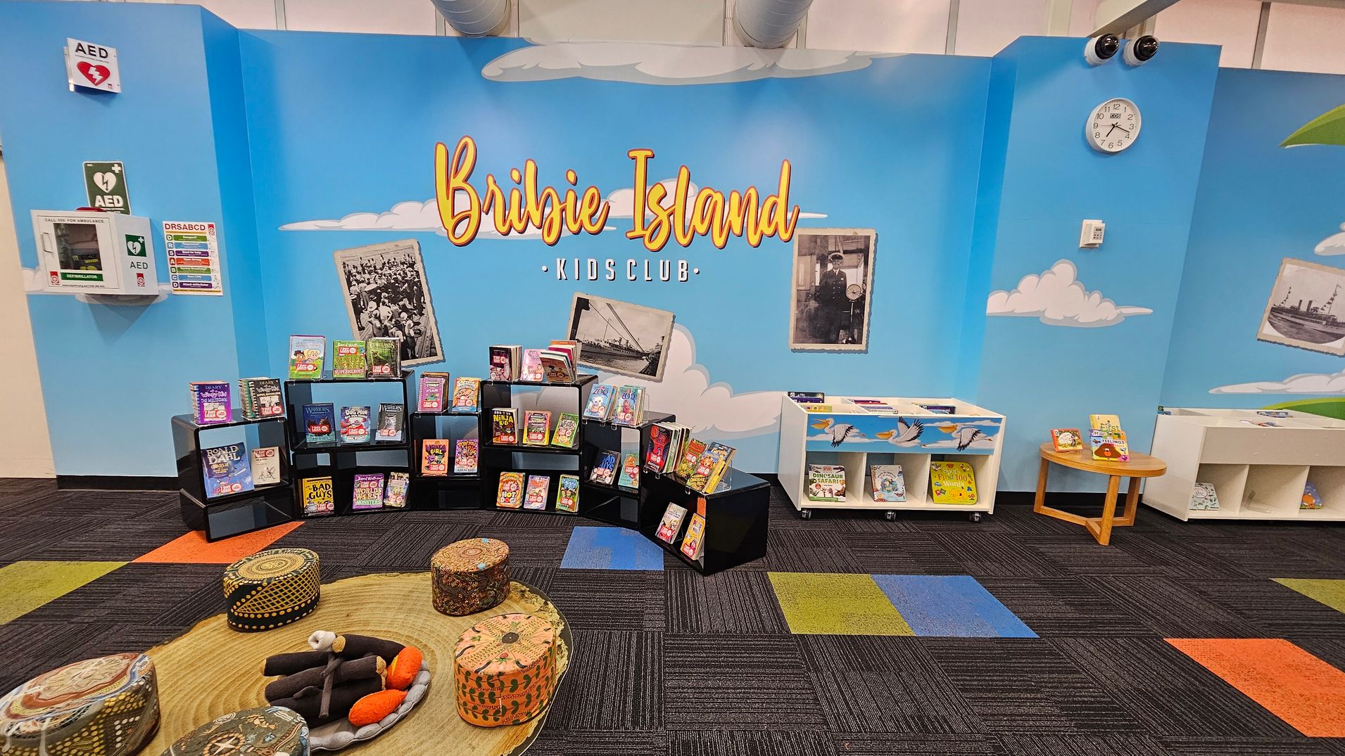 A children's reading area with a blue wall featuring the words "Bribie Island Kids Club" in large letters. Below, black shelves display various children's books. To the right, white shelves and a small round table hold more books. The floor is covered with a colourful carpet in shades of purple, green, blue, and orange. Cushioned stools with patterns are arranged around a circular mat. A clock and AED signs are mounted on the wall. Vintage photos are also displayed on the wall. A children's reading area with a blue wall featuring the words "Bribie Island Kids Club" in large letters. Below, black shelves display various children's books. To the right, white shelves and a small round table hold more books. The floor is covered with a colourful carpet in shades of purple, green, blue, and orange. Cushioned stools with patterns are arranged around a circular mat. A clock and AED signs are mounted on the wall. Vintage photos are also displayed on the wall.