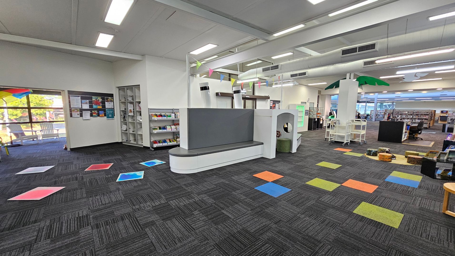 A library interior with a grey carpeted floor featuring colourful square tiles. On the left, a window with a view outside and a notice board with various papers. A bookshelf with books and magazines is nearby. In the centre, a white and grey seating area with a play structure. On the right, a palm tree decoration, book carts, and baskets on the floor. The ceiling has fluorescent lights and exposed ducts. A library interior with a grey carpeted floor featuring colourful square tiles. On the left, a window with a view outside and a notice board with various papers. A bookshelf with books and magazines is nearby. In the centre, a white and grey seating area with a play structure. On the right, a palm tree decoration, book carts, and baskets on the floor. The ceiling has fluorescent lights and exposed ducts.