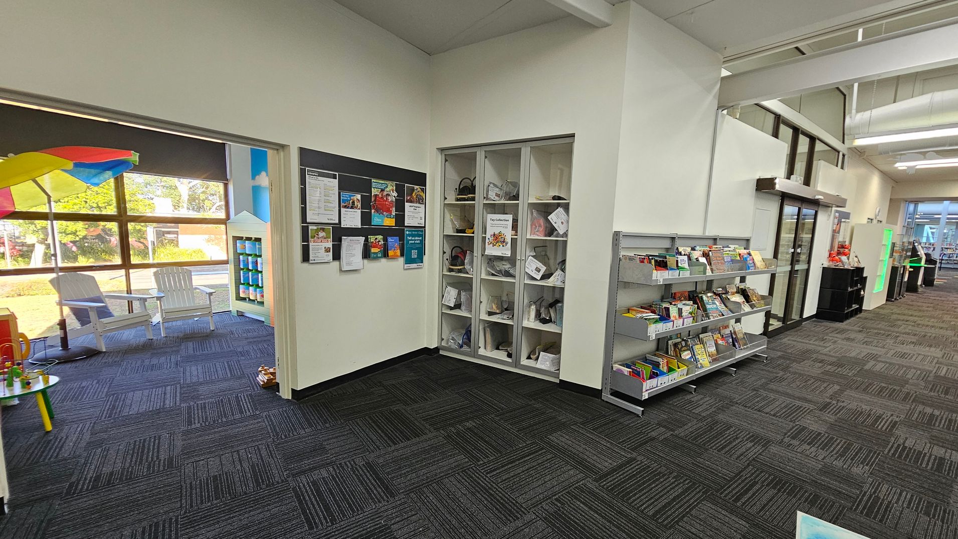 On the left, a room with large windows, white chairs, and a colourful umbrella. A small table with toys is visible. In the centre, a wall with a bulletin board displaying various notices and a shelf with toy collections. On the right, a bookshelf filled with books and a glass door leading to another area. The floor is covered with dark patterned carpet tiles, and the ceiling has exposed beams and lighting fixtures. On the left, a room with large windows, white chairs, and a colourful umbrella. A small table with toys is visible. In the centre, a wall with a bulletin board displaying various notices and a shelf with toy collections. On the right, a bookshelf filled with books and a glass door leading to another area. The floor is covered with dark patterned carpet tiles, and the ceiling has exposed beams and lighting fixtures.