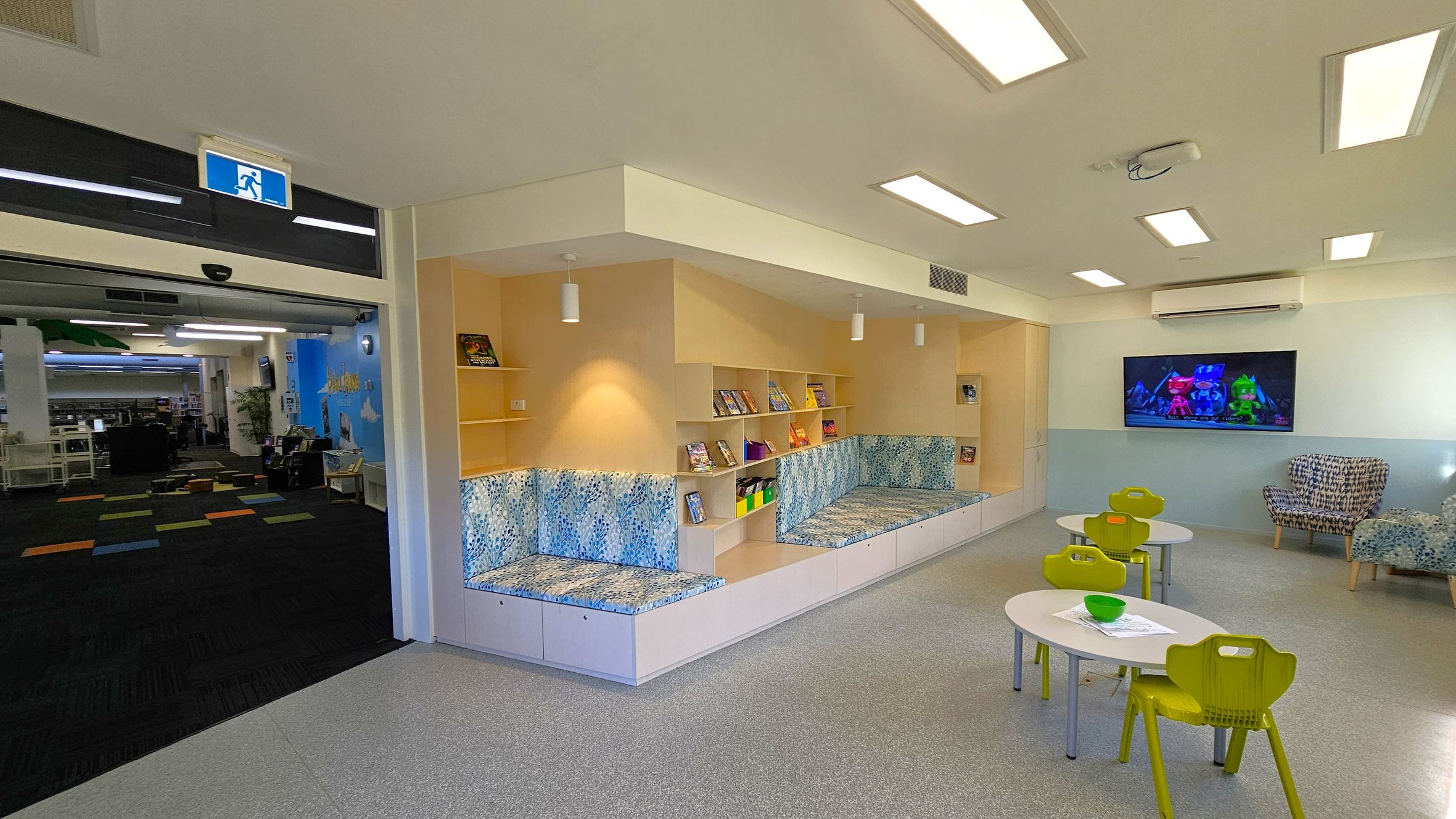 A children's reading area with a light grey floor and a ceiling with rectangular lights. On the left, there is an open entrance with a blue exit sign above, leading to a room with colourful carpet tiles. In the centre, there are built-in bookshelves with books and cushioned seating with a blue pattern. On the right, a wall-mounted TV displays an animated show above two small tables with green chairs. A patterned armchair is nearby. A children's reading area with a light grey floor and a ceiling with rectangular lights. On the left, there is an open entrance with a blue exit sign above, leading to a room with colourful carpet tiles. In the centre, there are built-in bookshelves with books and cushioned seating with a blue pattern. On the right, a wall-mounted TV displays an animated show above two small tables with green chairs. A patterned armchair is nearby.