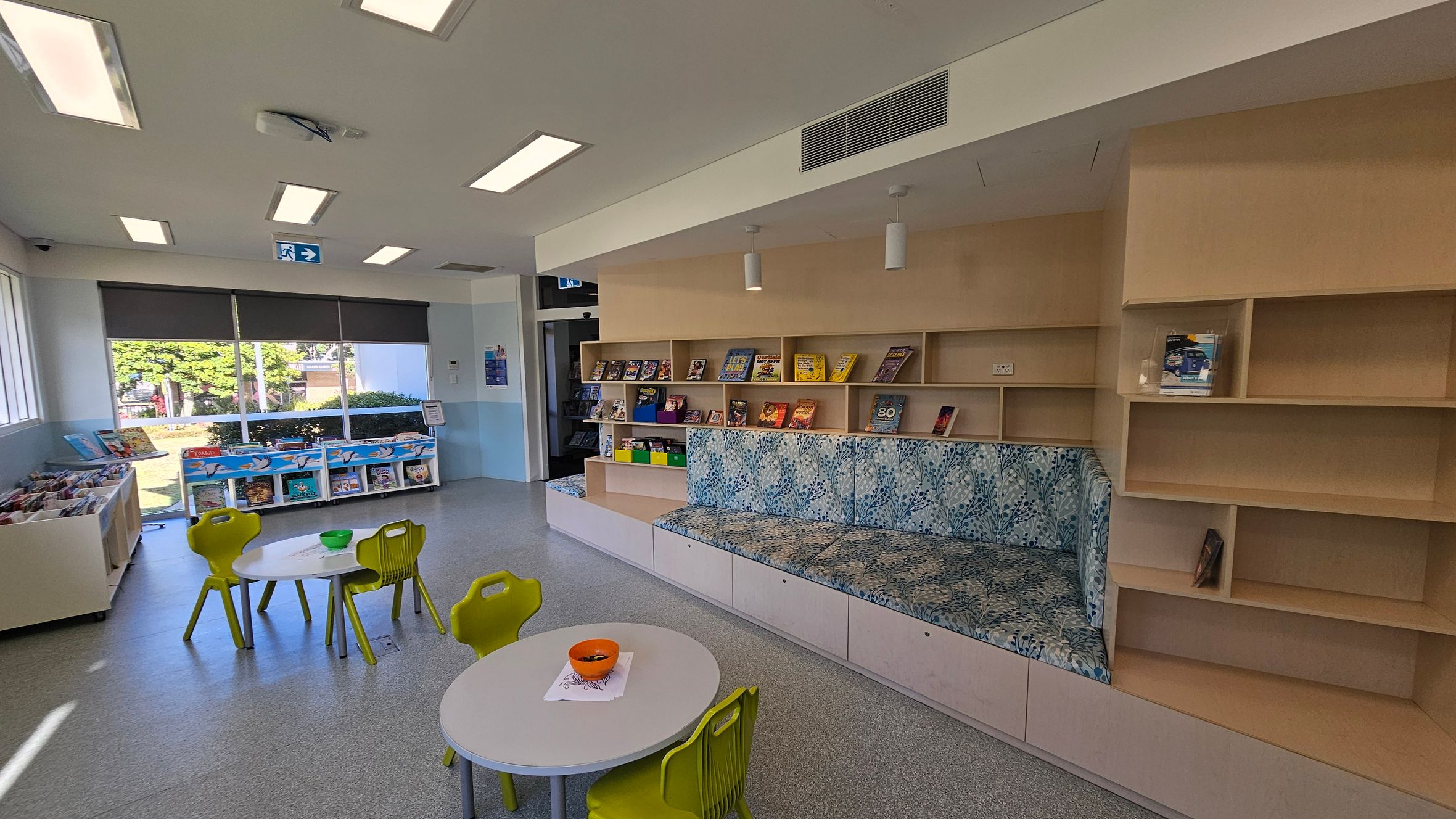 On the left, there are large windows with a view of trees and a building outside. Below the windows, there are shelves filled with books. In the centre, two small round tables with green chairs are placed on a grey floor, with a bowl on one table. On the right, there is a built-in wooden bookshelf with books and a cushioned seating area with a patterned fabric. The ceiling has rectangular lights and an air vent. On the left, there are large windows with a view of trees and a building outside. Below the windows, there are shelves filled with books. In the centre, two small round tables with green chairs are placed on a grey floor, with a bowl on one table. On the right, there is a built-in wooden bookshelf with books and a cushioned seating area with a patterned fabric. The ceiling has rectangular lights and an air vent.
