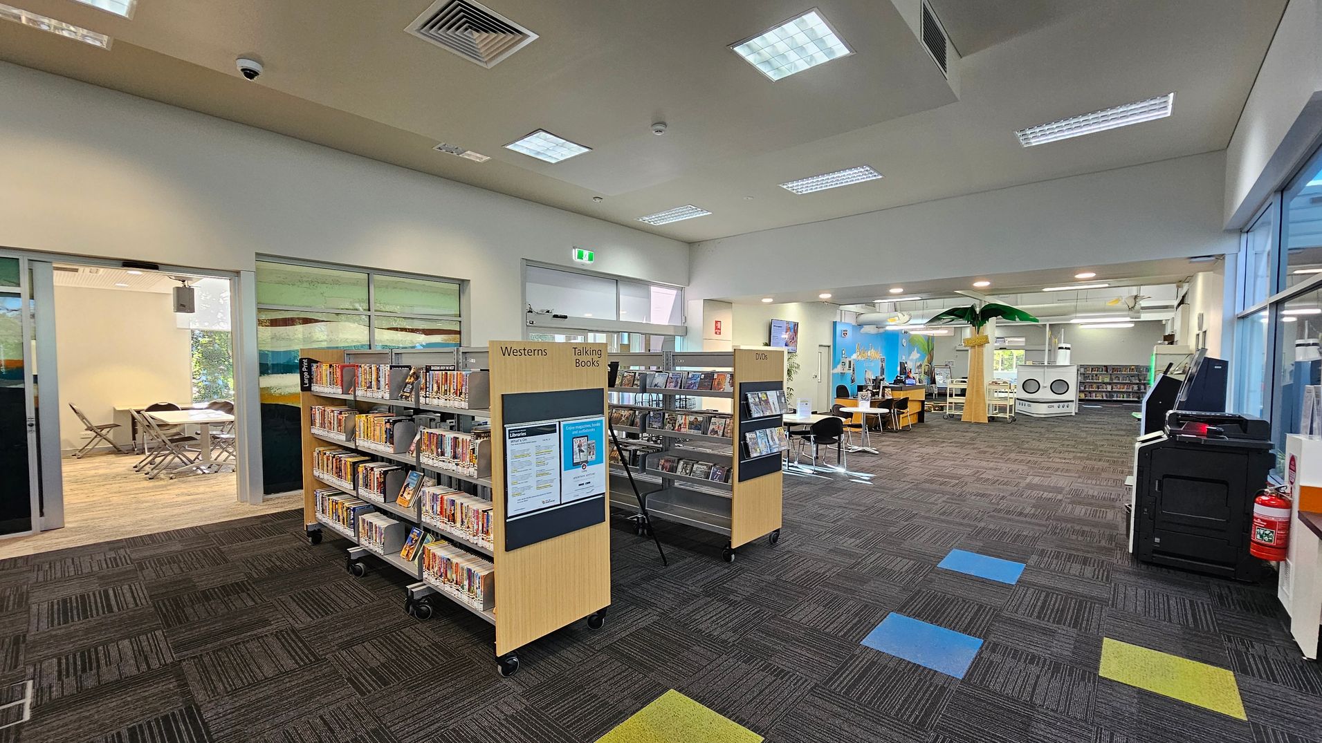 A library interior with a patterned carpet floor featuring blue and yellow squares. On the left, there are bookshelves labelled "Westerns" and "Talking Books," with DVDs on the right. In the background, a room with tables and chairs is visible through glass doors. The right side has a large palm tree decoration and a seating area with tables and chairs. The ceiling has recessed lighting and air vents. A fire extinguisher and a black machine are on the far right. A library interior with a patterned carpet floor featuring blue and yellow squares. On the left, there are bookshelves labelled "Westerns" and "Talking Books," with DVDs on the right. In the background, a room with tables and chairs is visible through glass doors. The right side has a large palm tree decoration and a seating area with tables and chairs. The ceiling has recessed lighting and air vents. A fire extinguisher and a black machine are on the far right.
